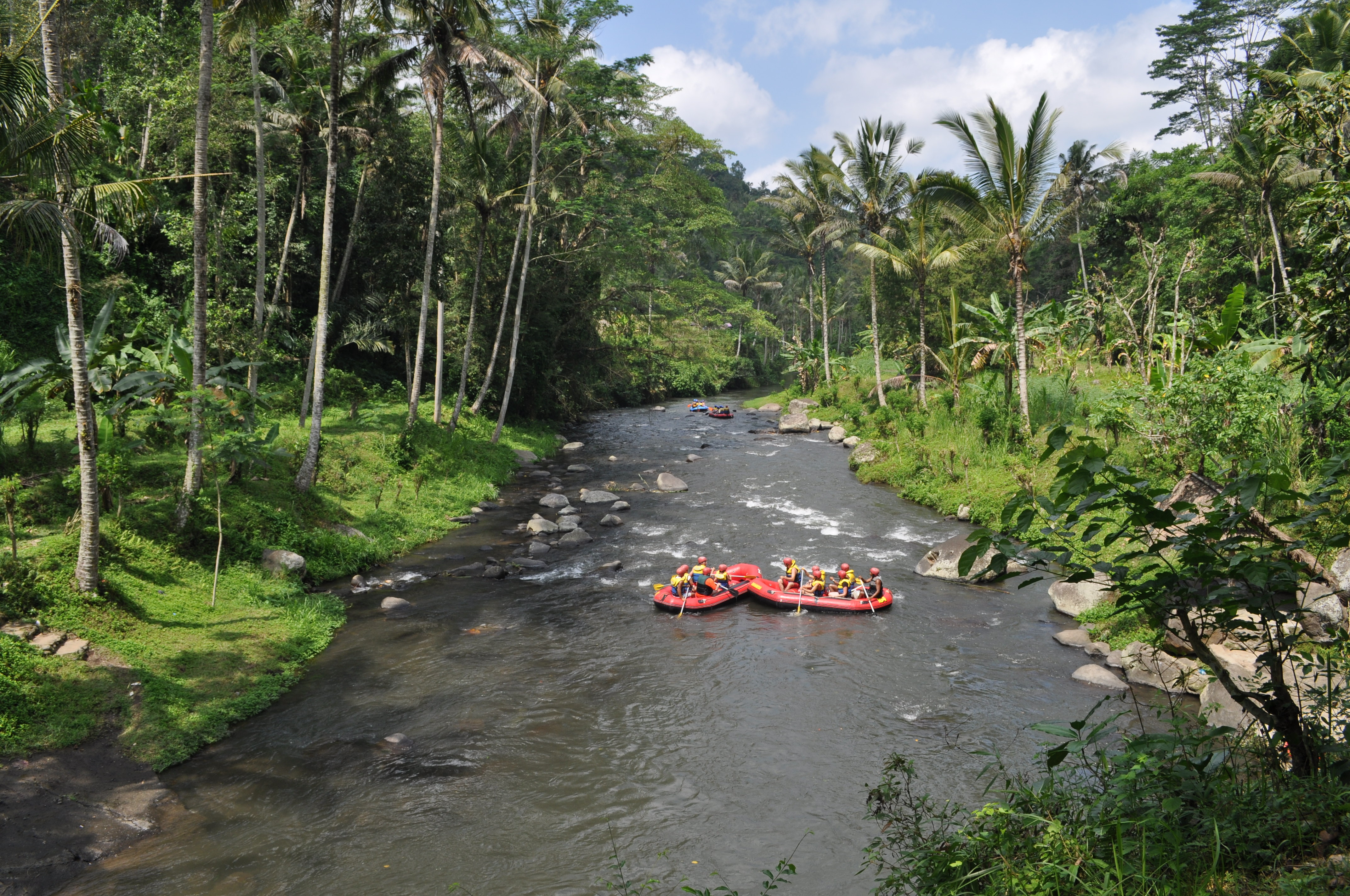 Two rafting boats float down a palm-lined Bali river, surrounded by boulders, jungle trees, and bright green banks.