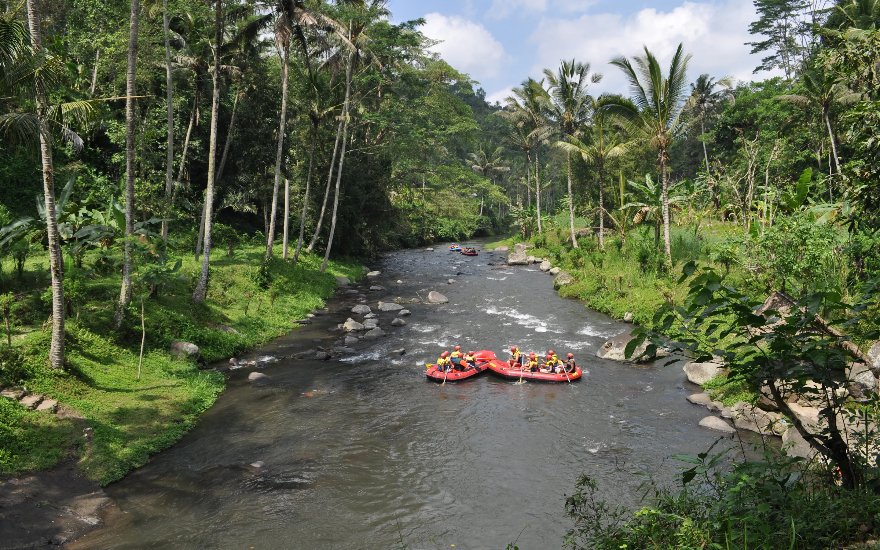 Two rafting boats float down a palm-lined Bali river, surrounded by boulders, jungle trees, and bright green banks.