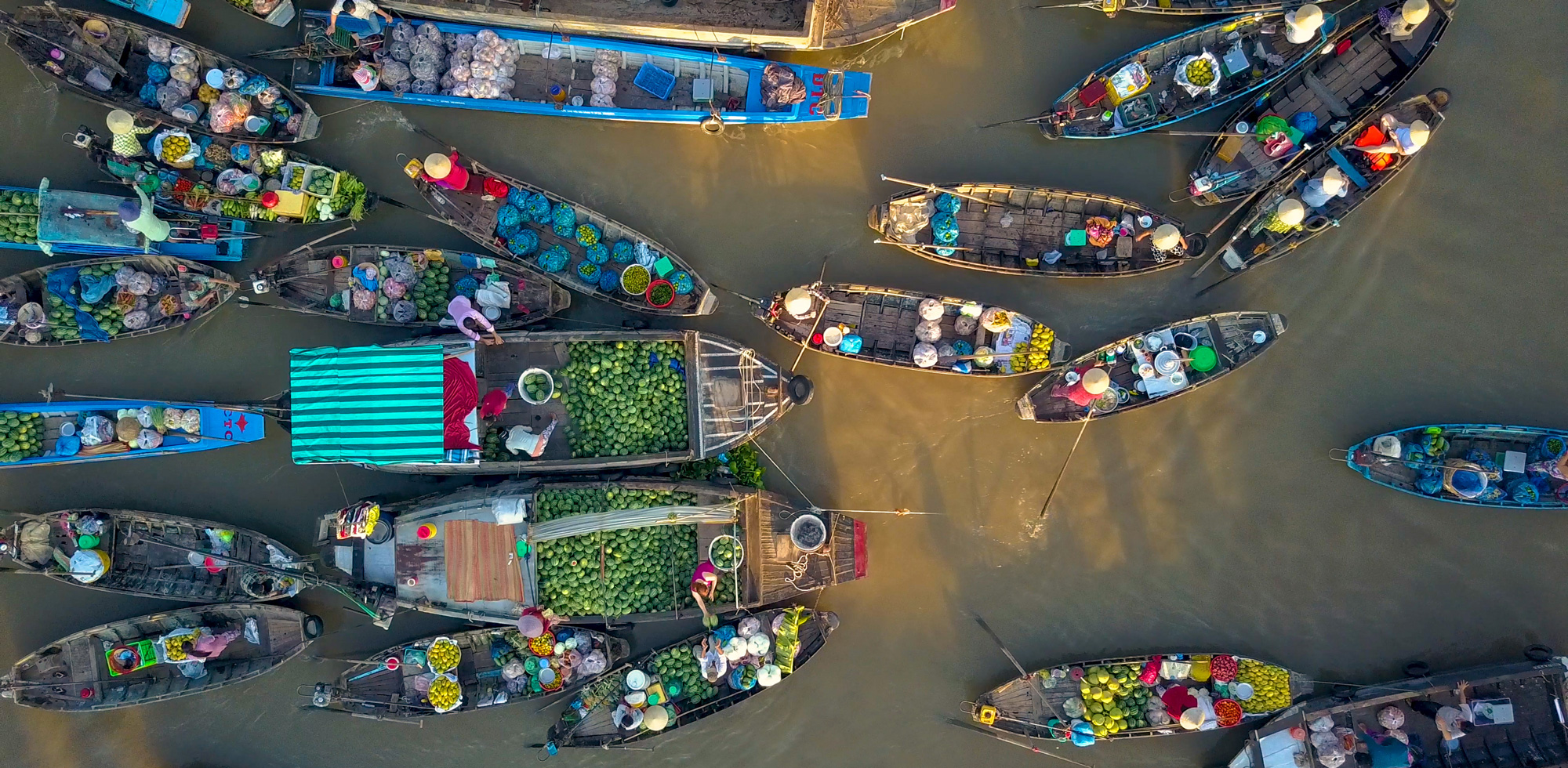 An aerial view shows colorful boats clustered in a Mekong Delta canal, their cargoes packed tightly along muddy water.