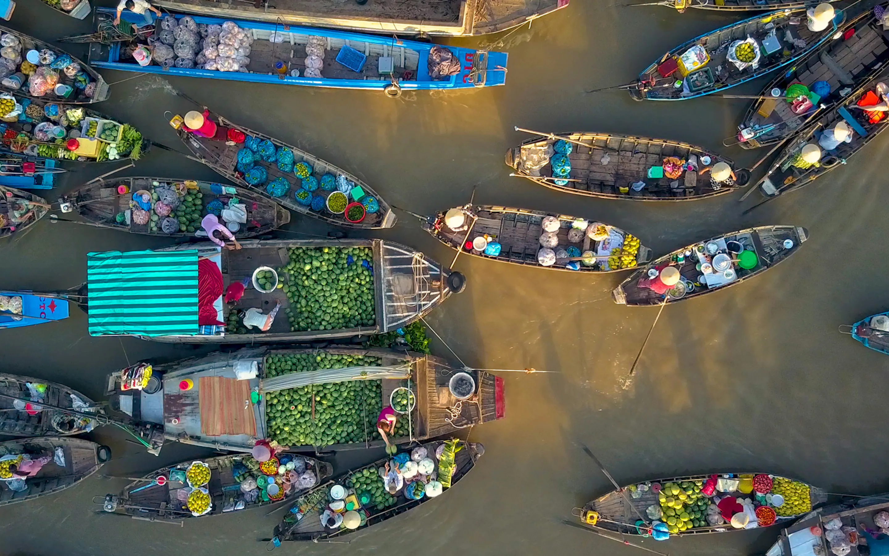 An aerial view shows colorful boats clustered in a Mekong Delta canal, their cargoes packed tightly along muddy water.