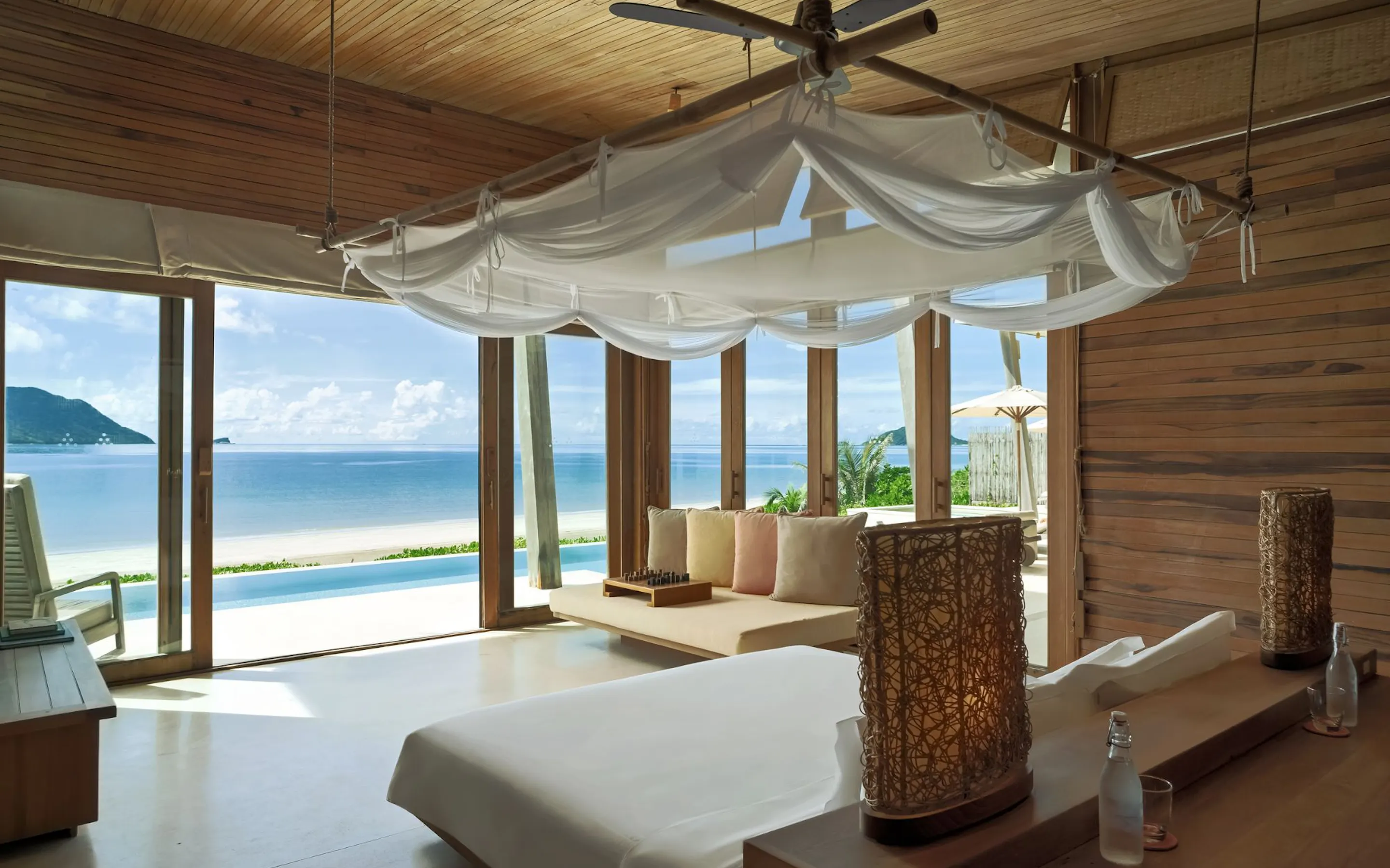 A wood-paneled bedroom opens to a private pool and ocean view at Six Senses Con Dao, framed by airy white drapes.
