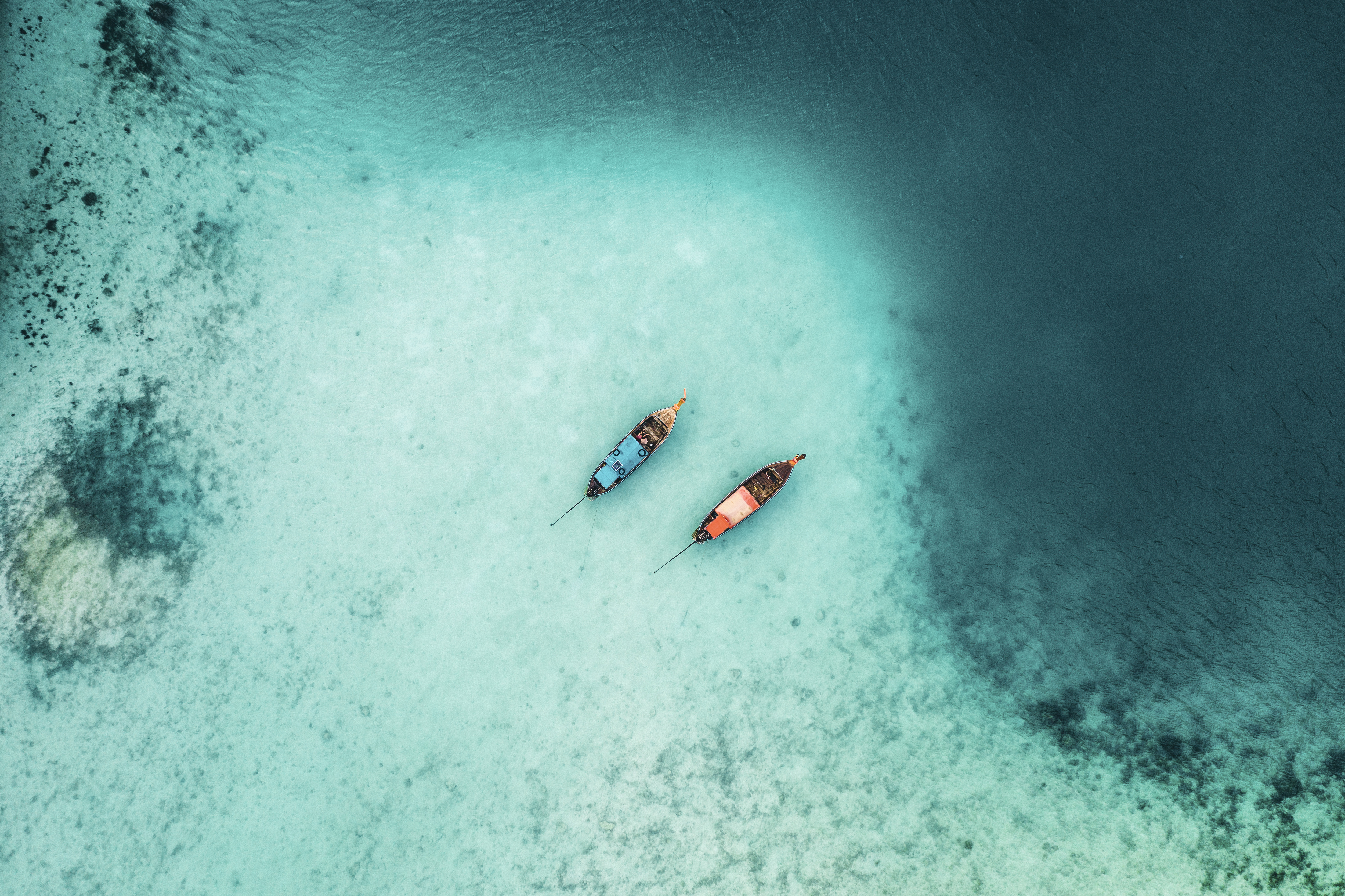 An aerial view shows two longtail boats drifting above pale sand and reef patterns in clear turquoise water.