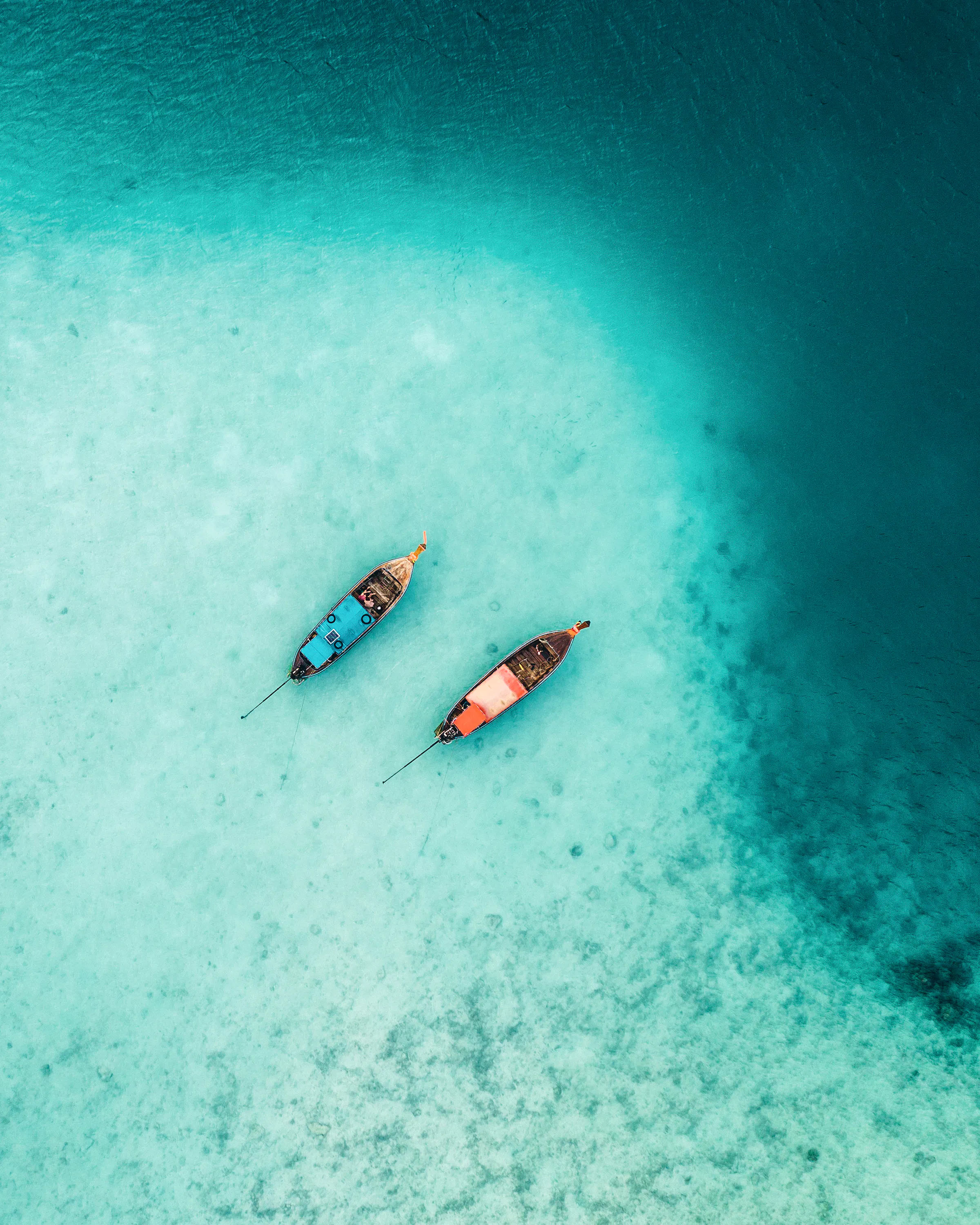 An aerial view shows two longtail boats drifting above pale sand and reef patterns in clear turquoise water.