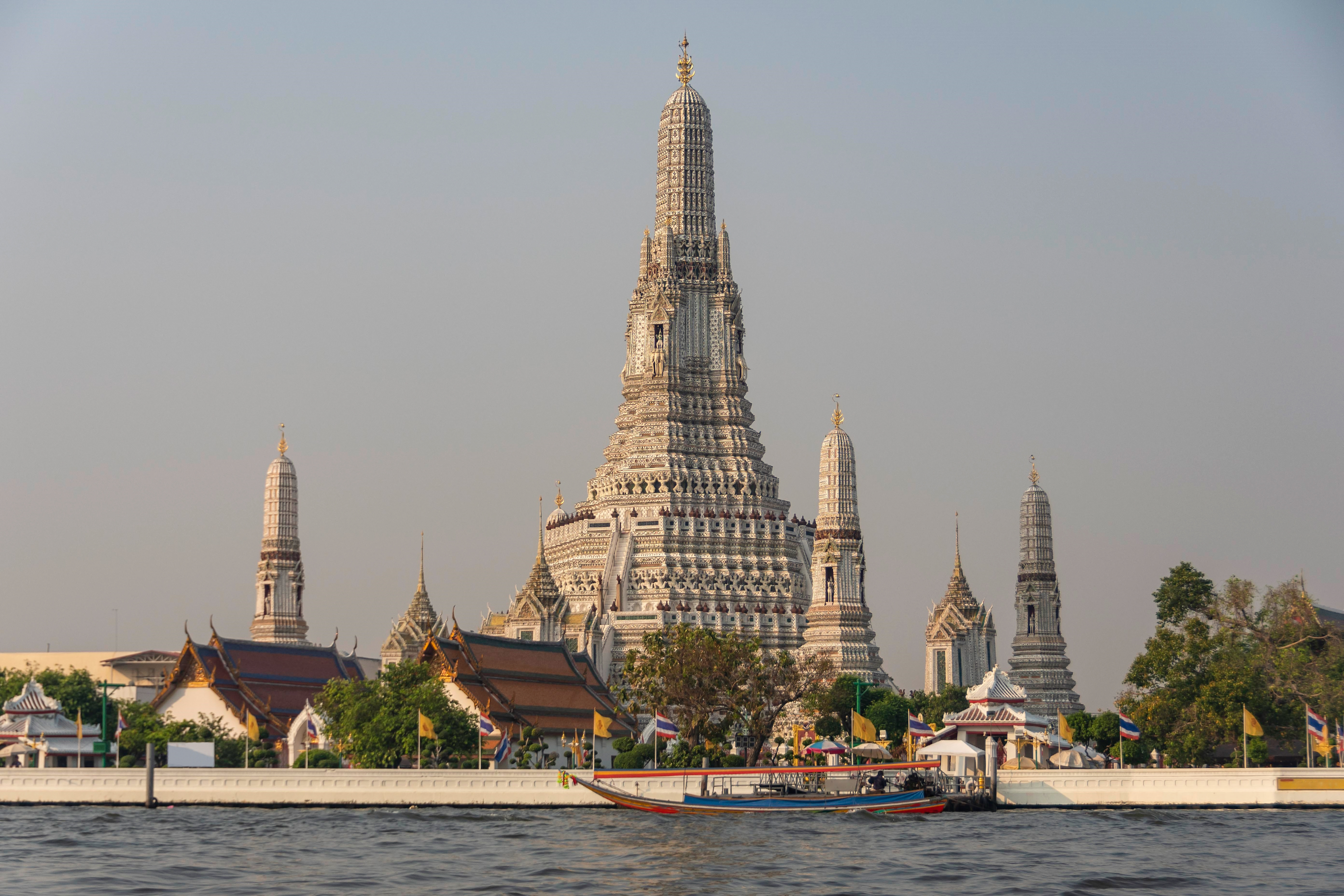 Wat Arun rises above the Chao Phraya River in Bangkok, where a long-tail boat passes the temple frontage.