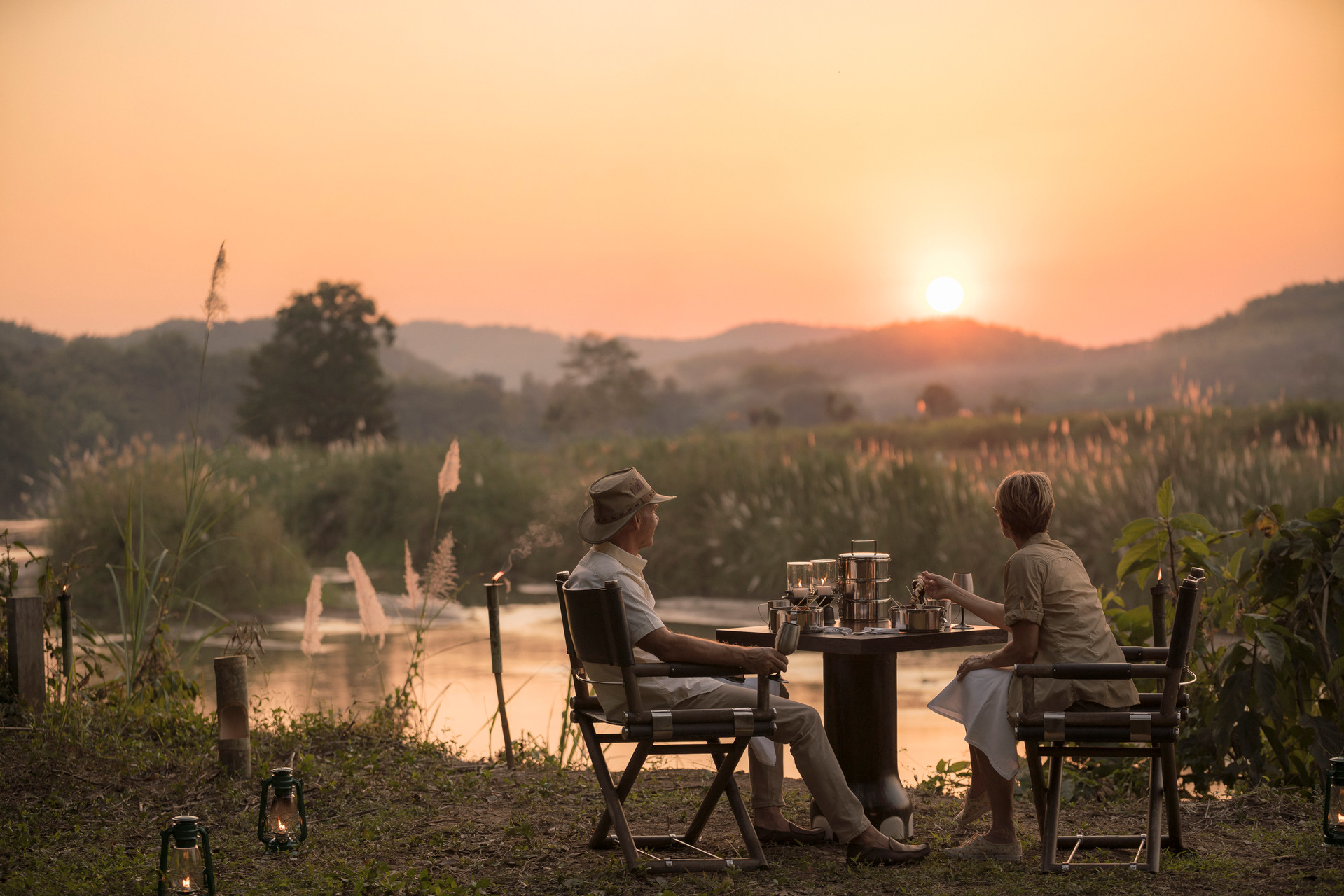 Two guests sit at a riverside table at sunset, with lanterns glowing softly and hazy hills beyond the water.