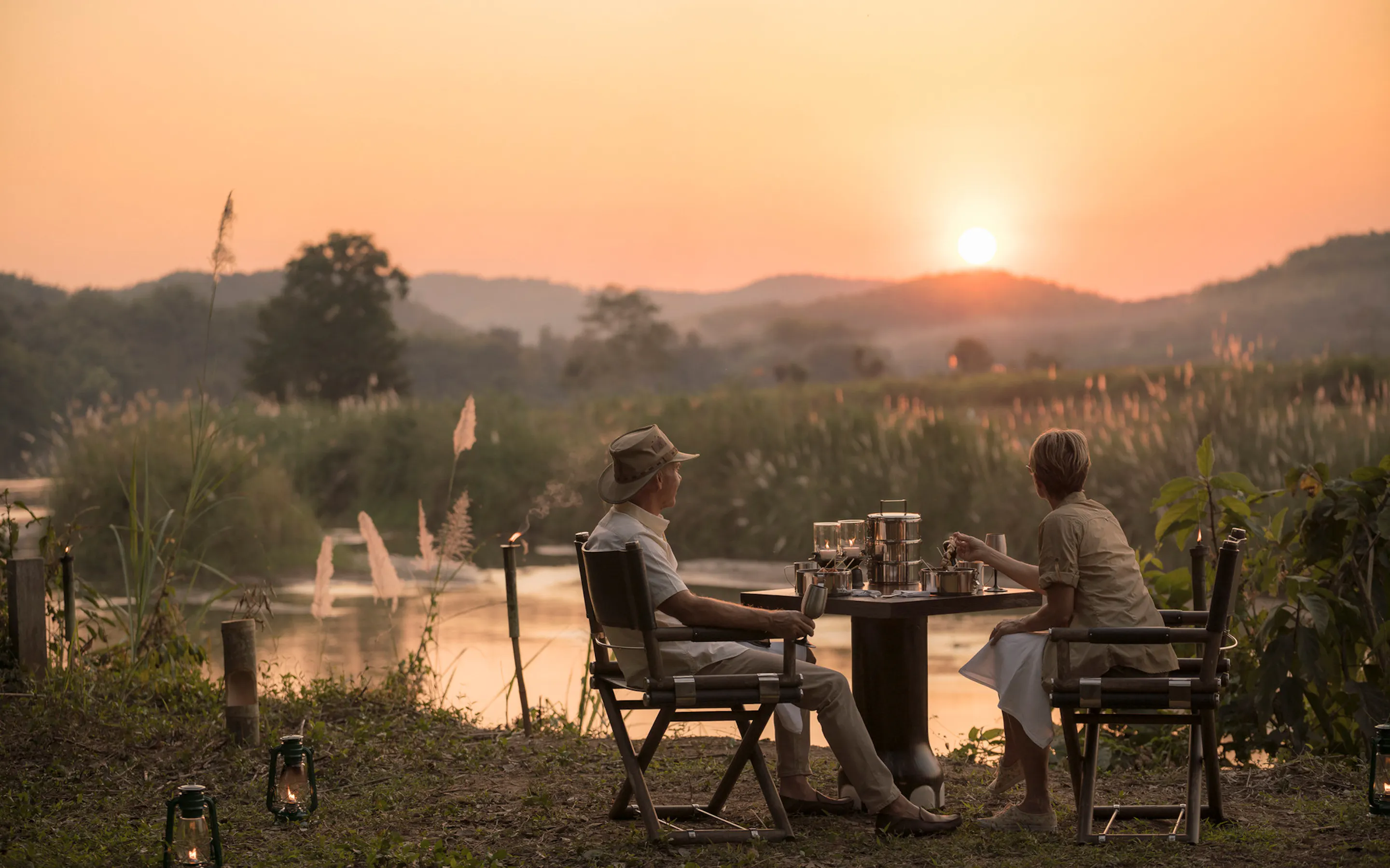 Two guests sit at a riverside table at sunset, with lanterns glowing softly and hazy hills beyond the water.