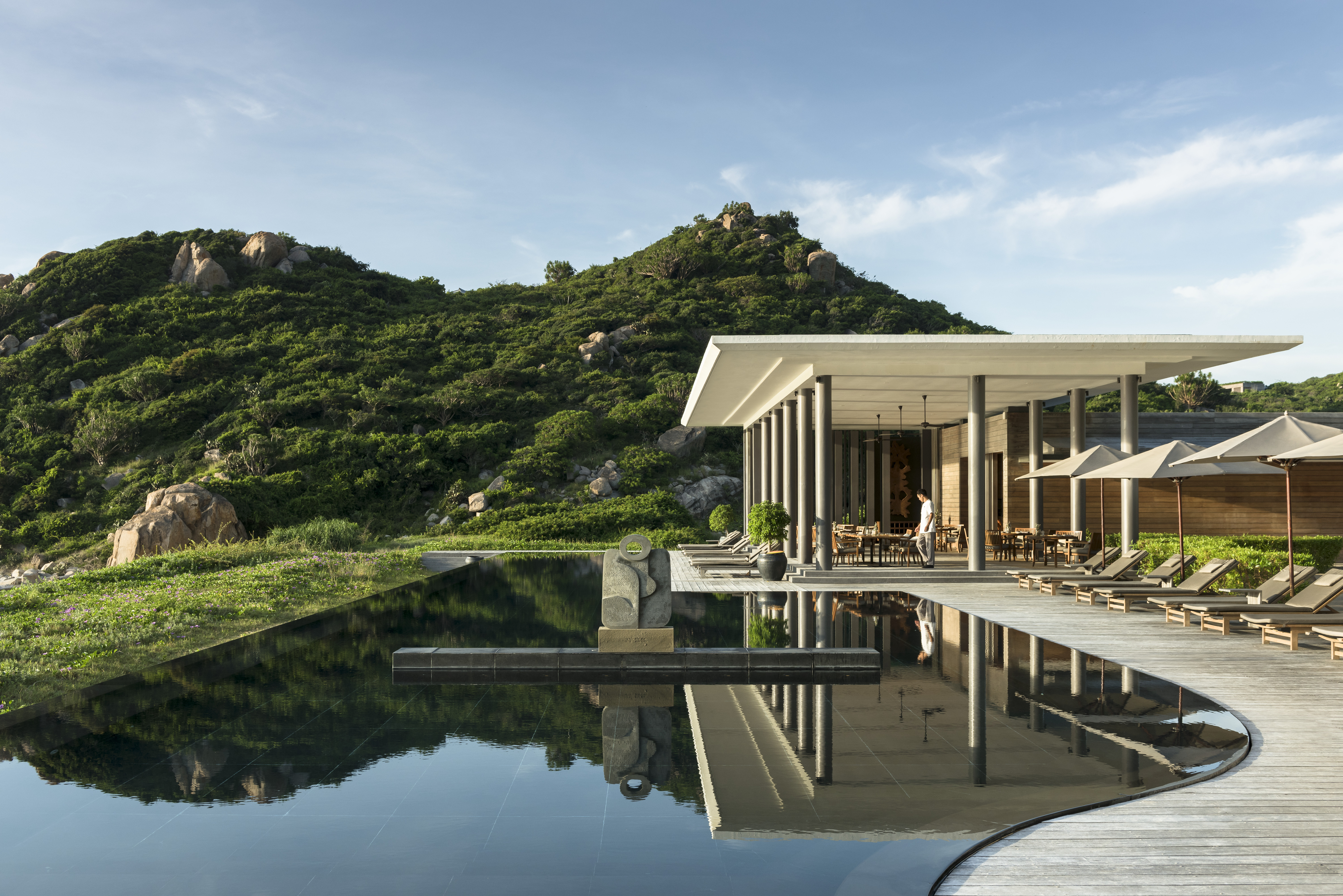 A curved reflecting pool mirrors Amanoi Beach Club, set between green hills and smooth boulders above the coast.