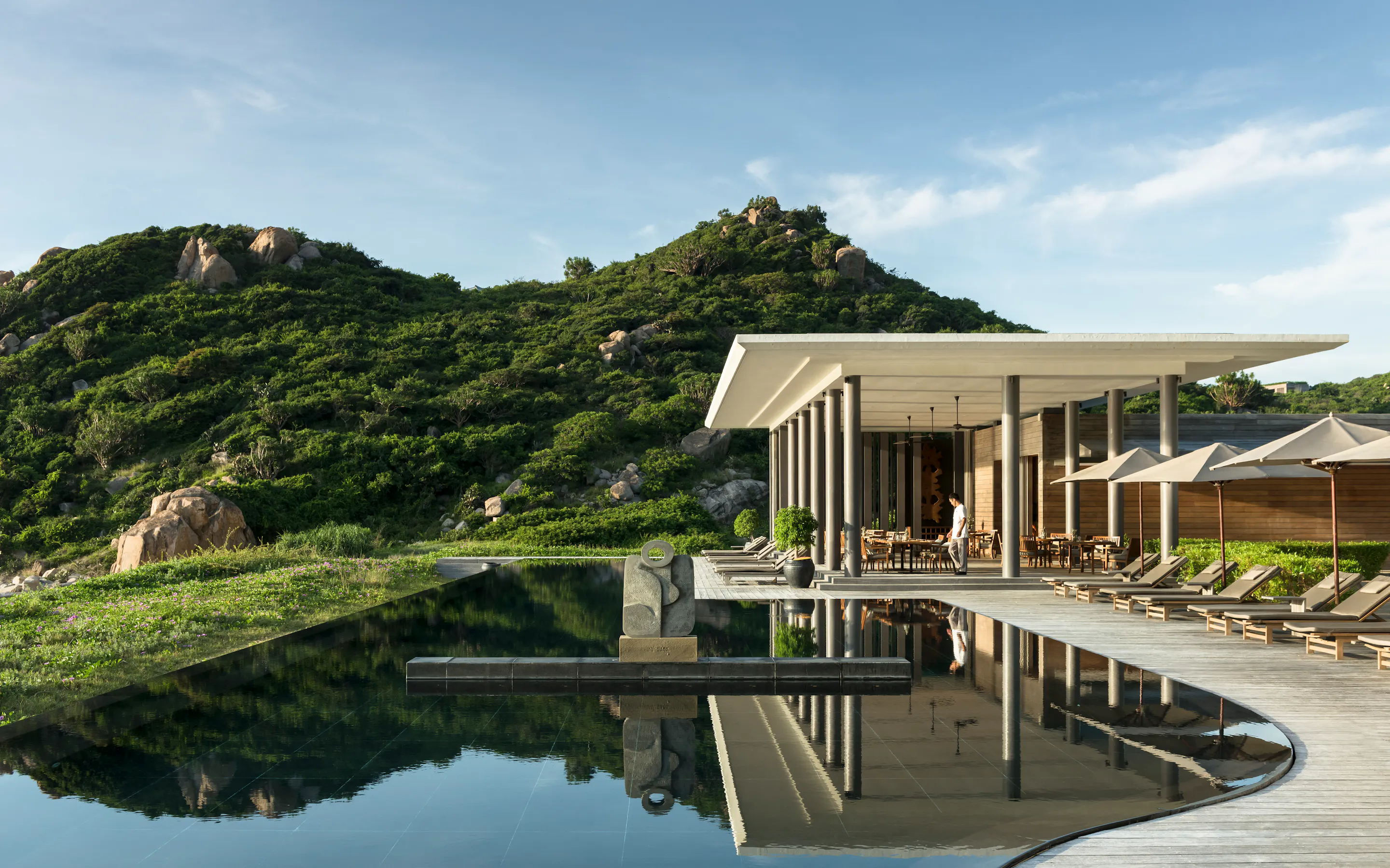 A curved reflecting pool mirrors Amanoi Beach Club, set between green hills and smooth boulders above the coast.