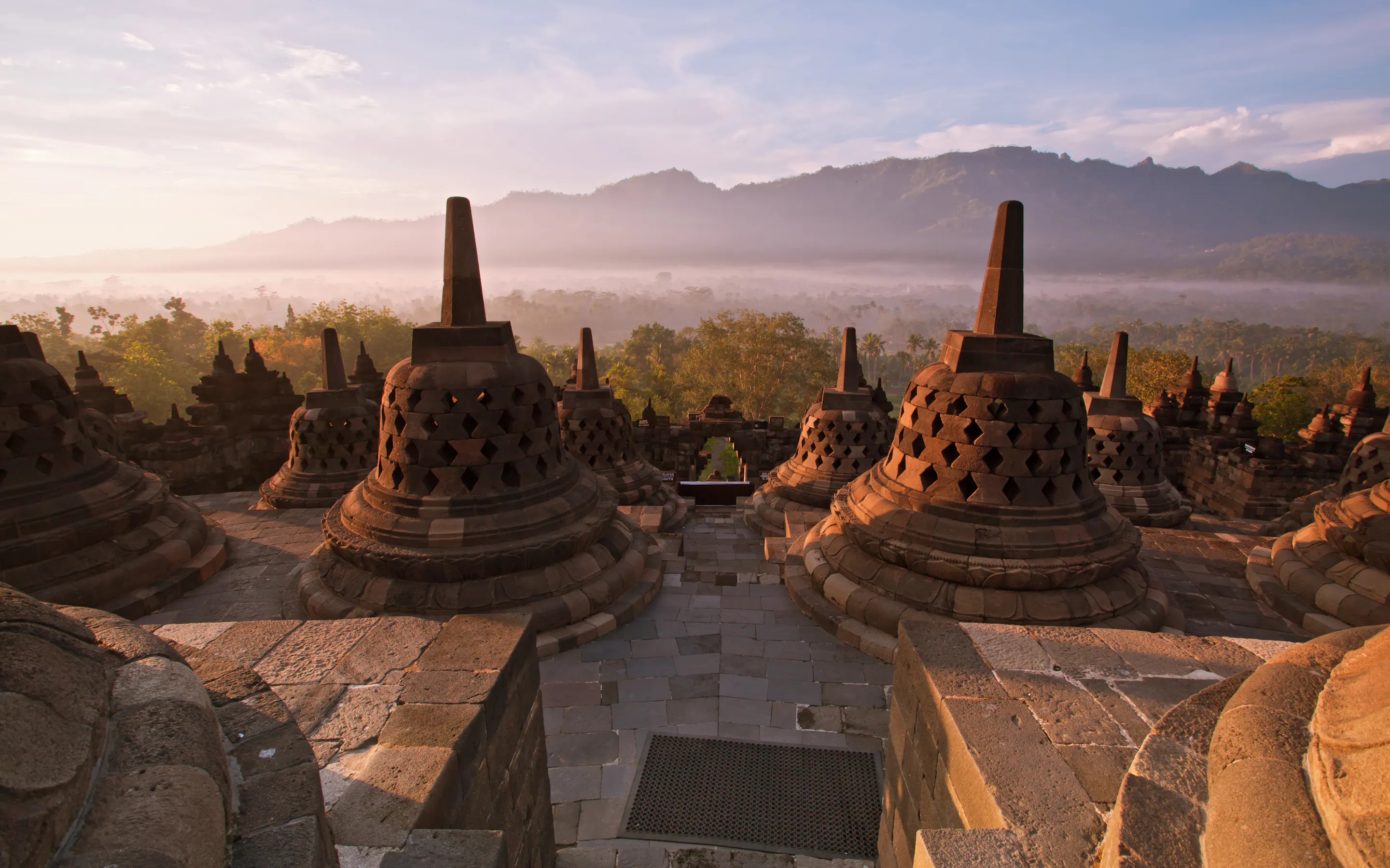 Borobudur's stone stupas catch the morning sun above a misty plain, with dark mountain ridges rising beyond them.