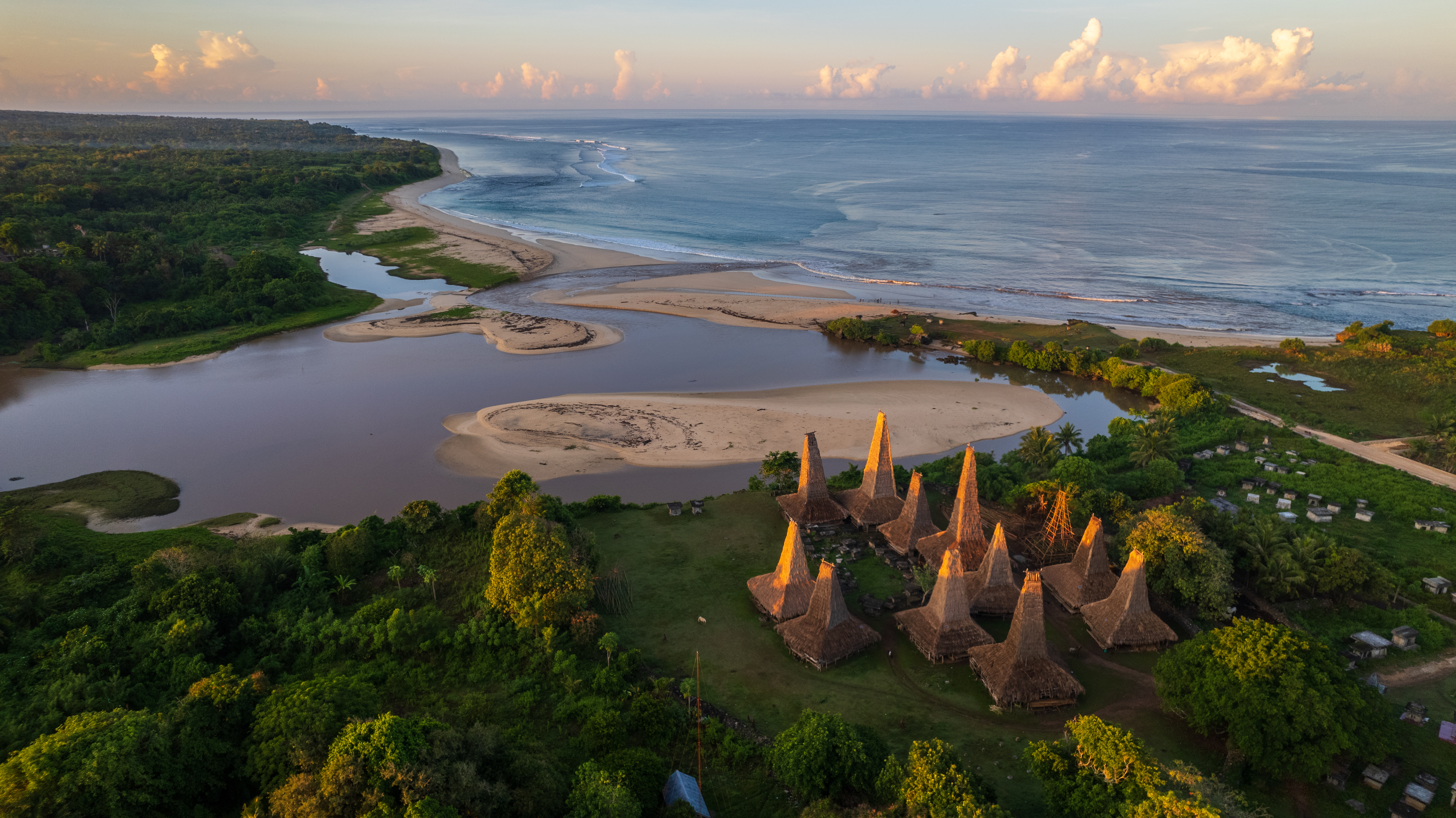 Tall thatched village roofs stand beside a winding estuary on Sumba, with sandbars and the sea stretching beyond.