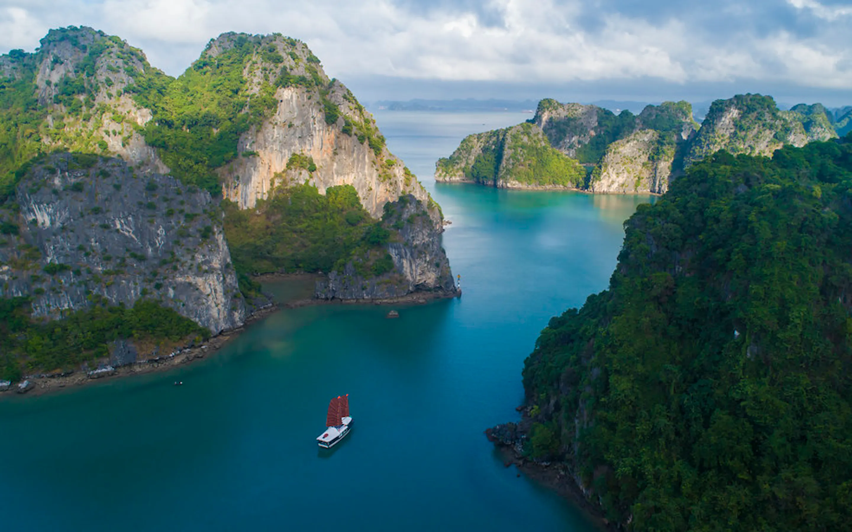 A red-sailed junk drifts through turquoise water in Ha Long Bay, framed by steep limestone islets and a cloudy sky.