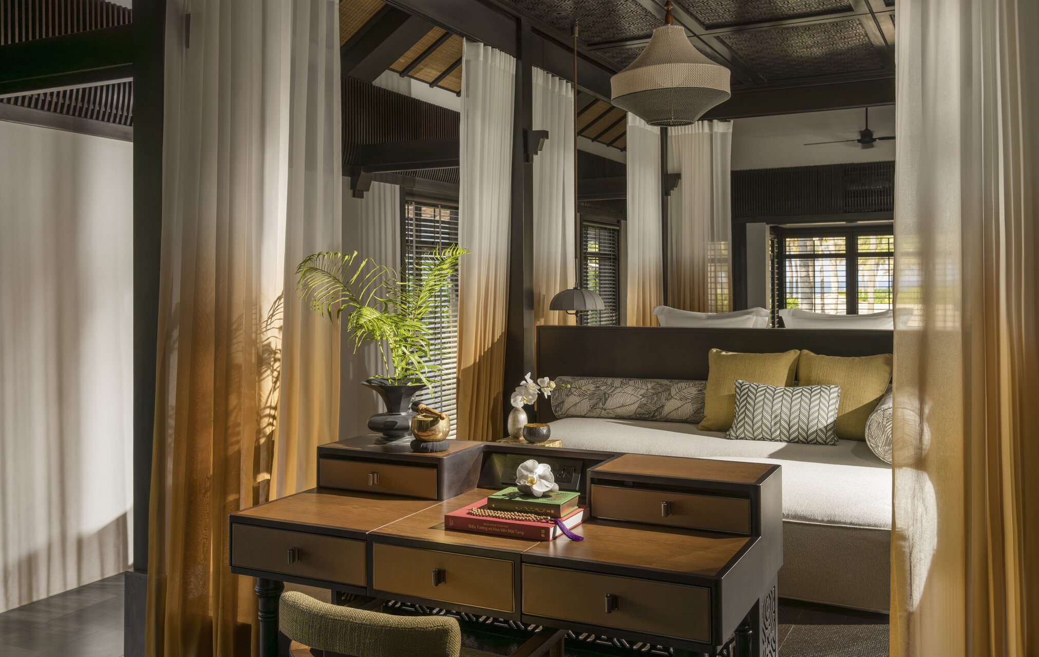 Sunlight falls through sheer curtains onto a daybed and desk inside a dark wood suite at Four Seasons The Nam Hai.