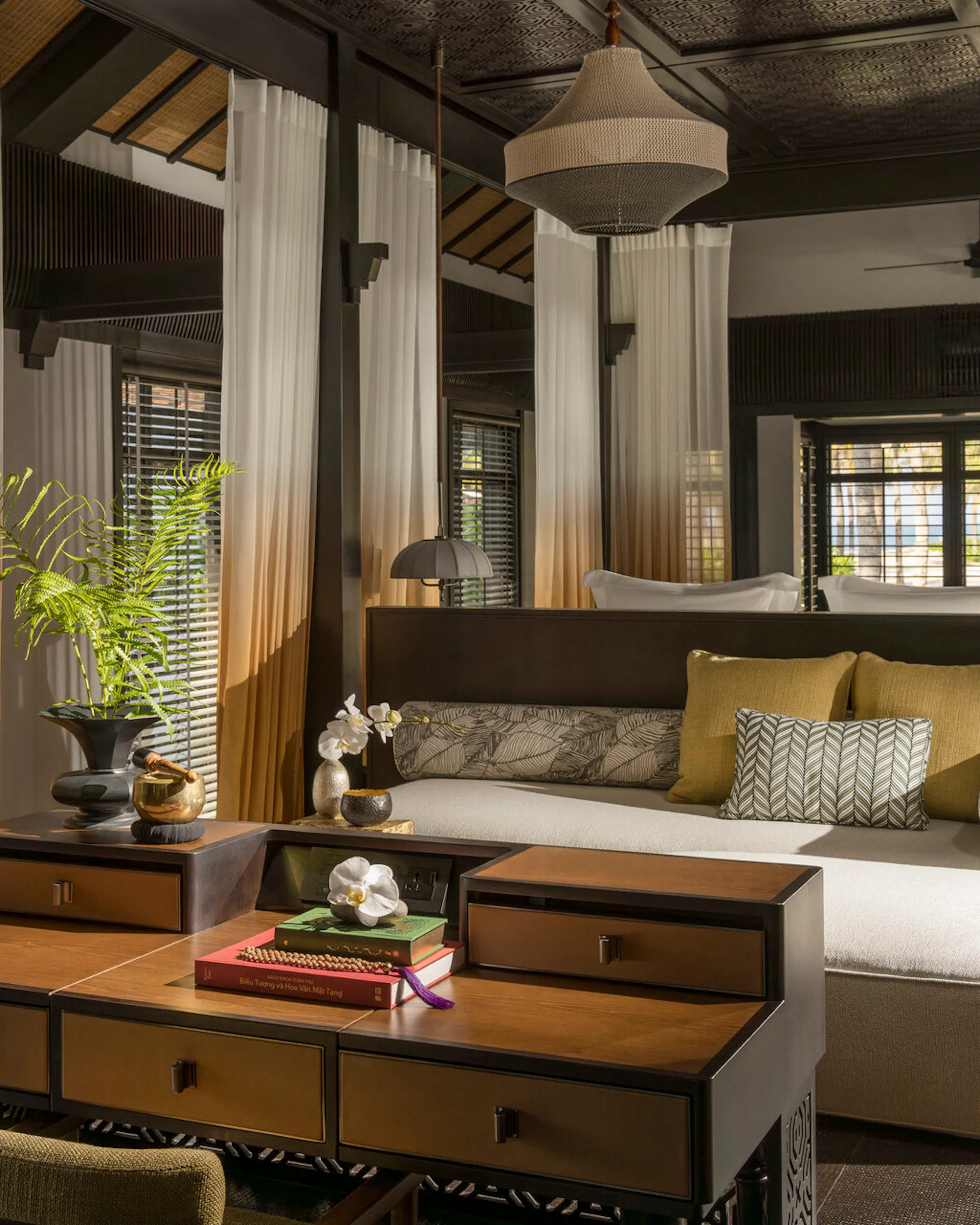 Sunlight falls through sheer curtains onto a daybed and desk inside a dark wood suite at Four Seasons The Nam Hai.