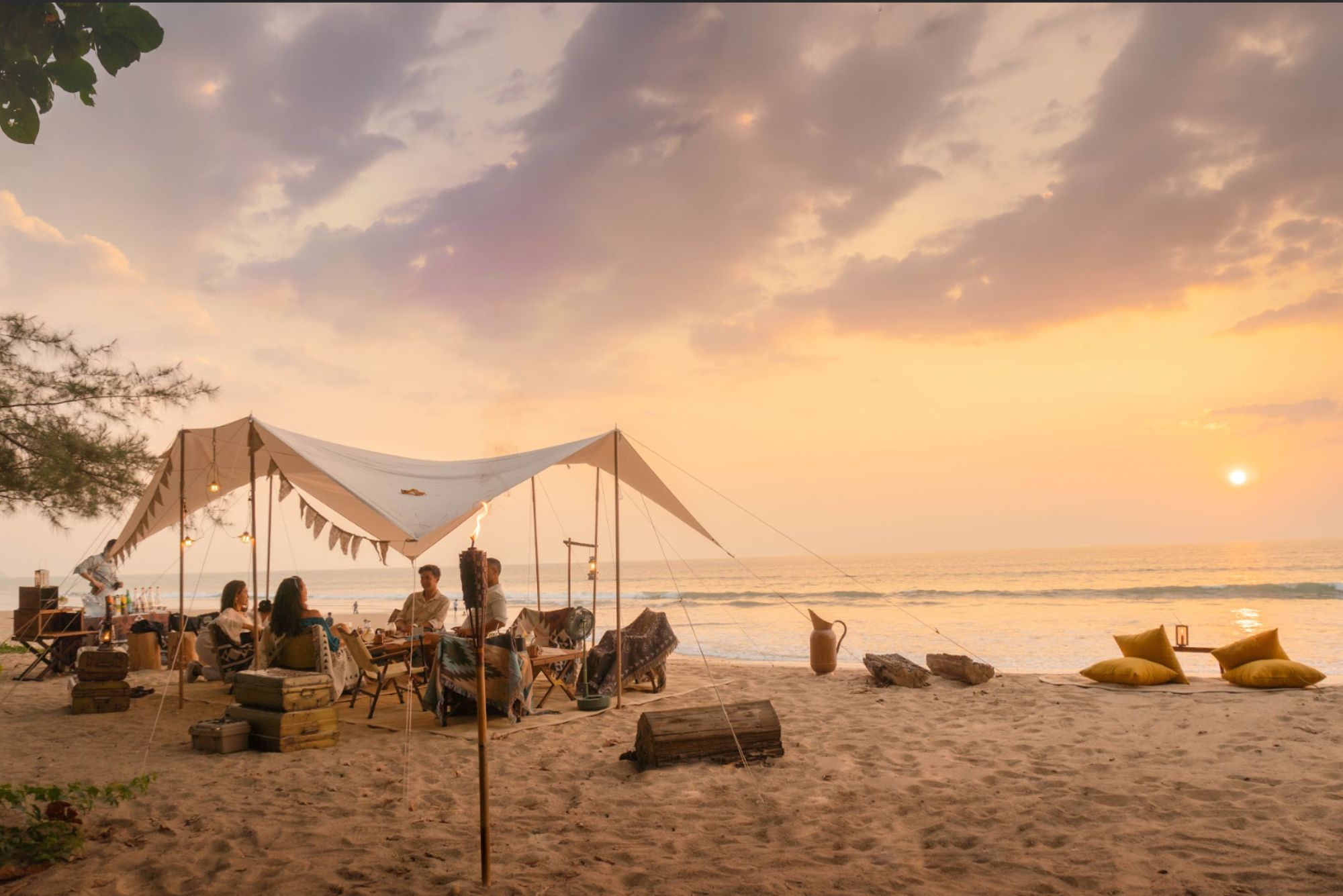 Guests gather beneath a canvas tent on Khao Lak beach, with pale sand, calm surf, and sunset stretching behind.