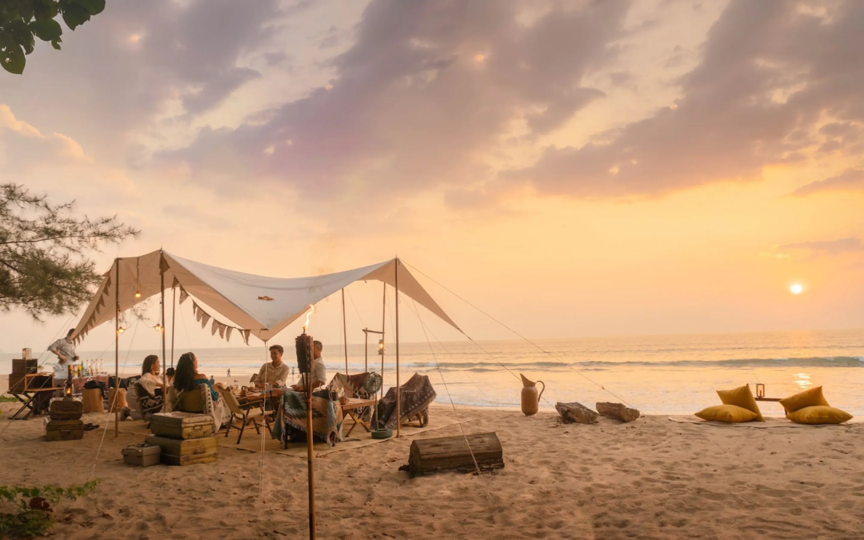 Guests gather beneath a canvas tent on Khao Lak beach, with pale sand, calm surf, and sunset stretching behind.