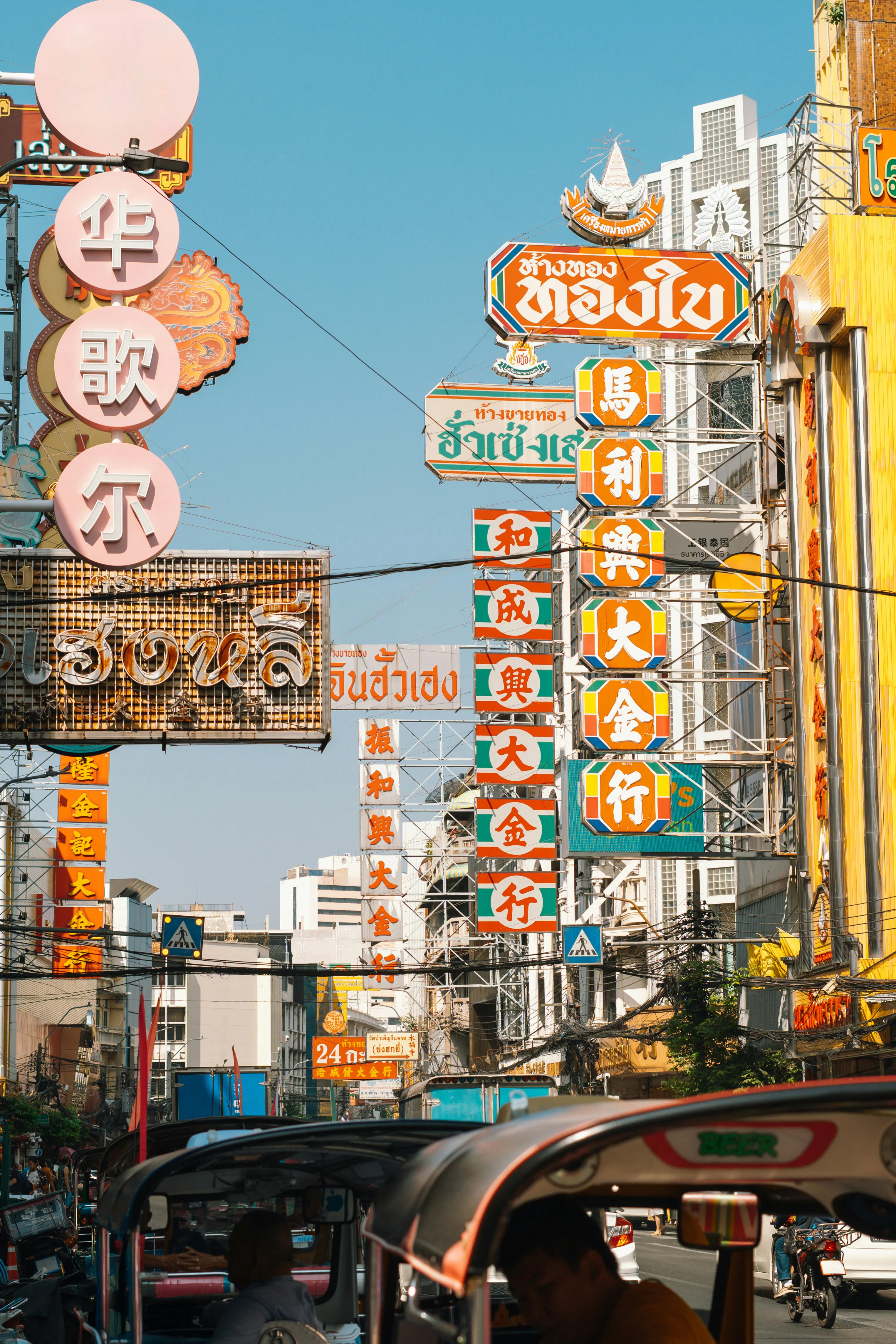 Colorful vertical signs rise above a busy Bangkok Chinatown street, with tuk-tuks, traffic and bright facades below.