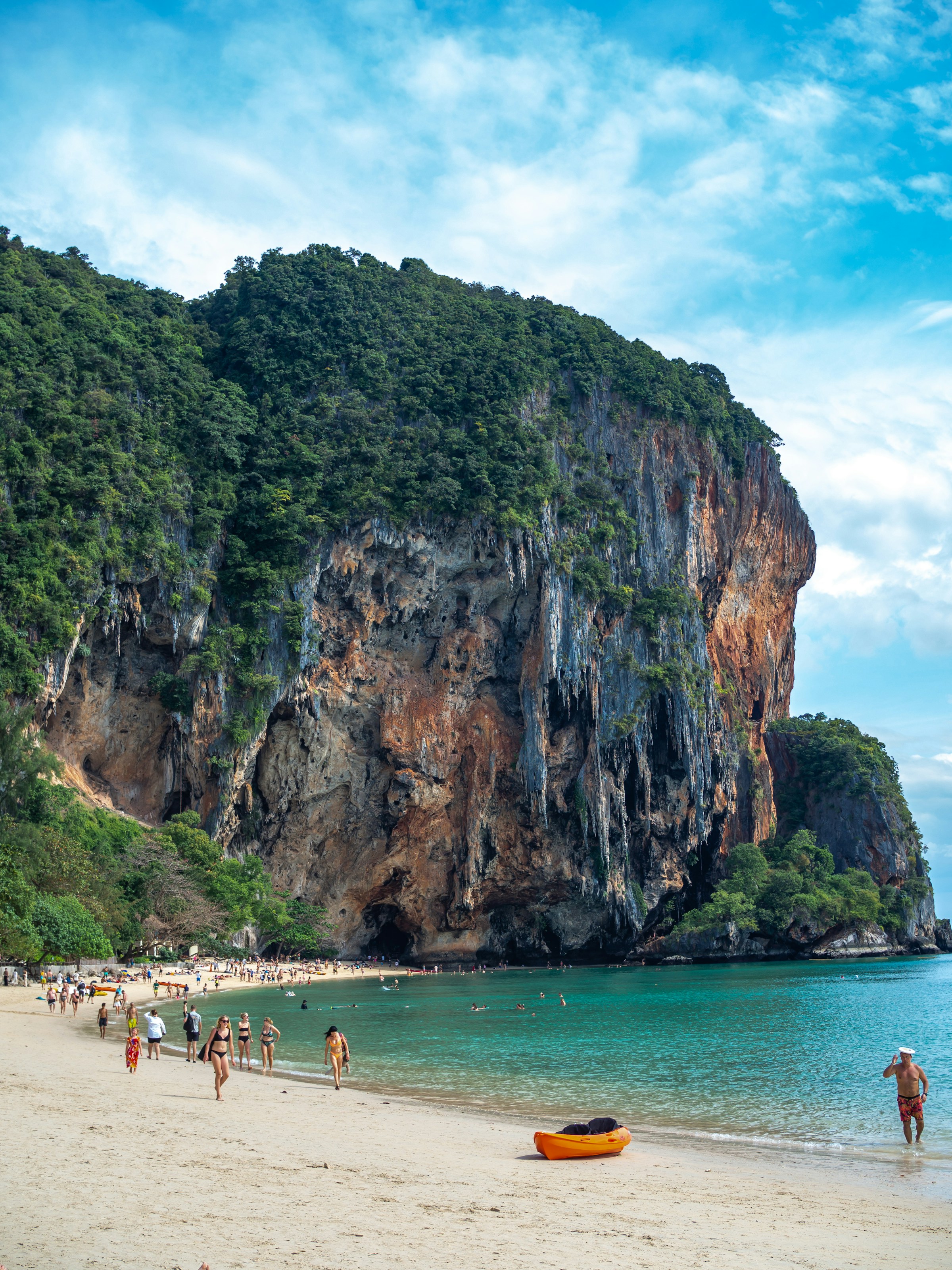 Beachgoers walk beside turquoise water under a towering limestone cliff, with a yellow kayak resting on the sand.