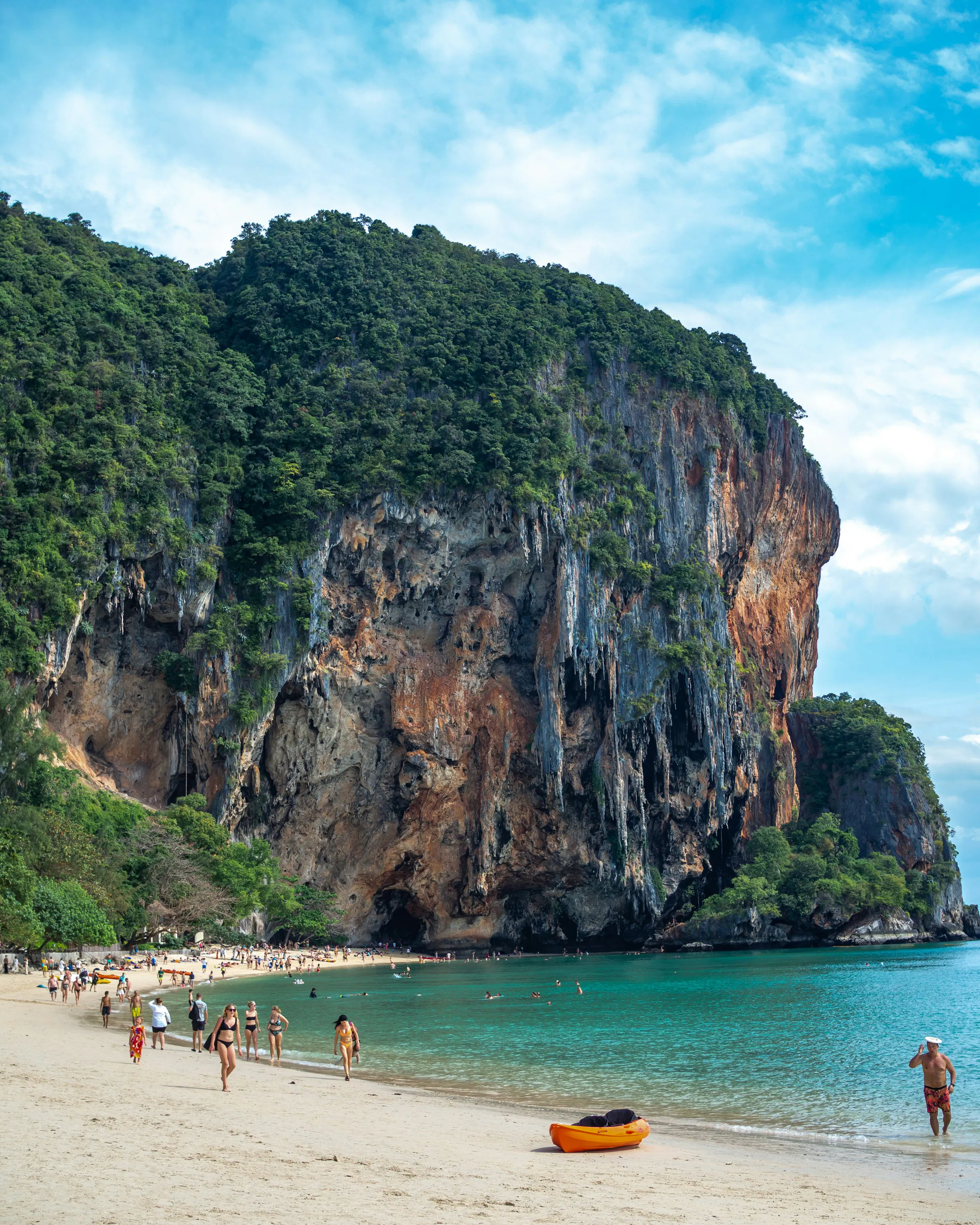 Beachgoers walk beside turquoise water under a towering limestone cliff, with a yellow kayak resting on the sand.