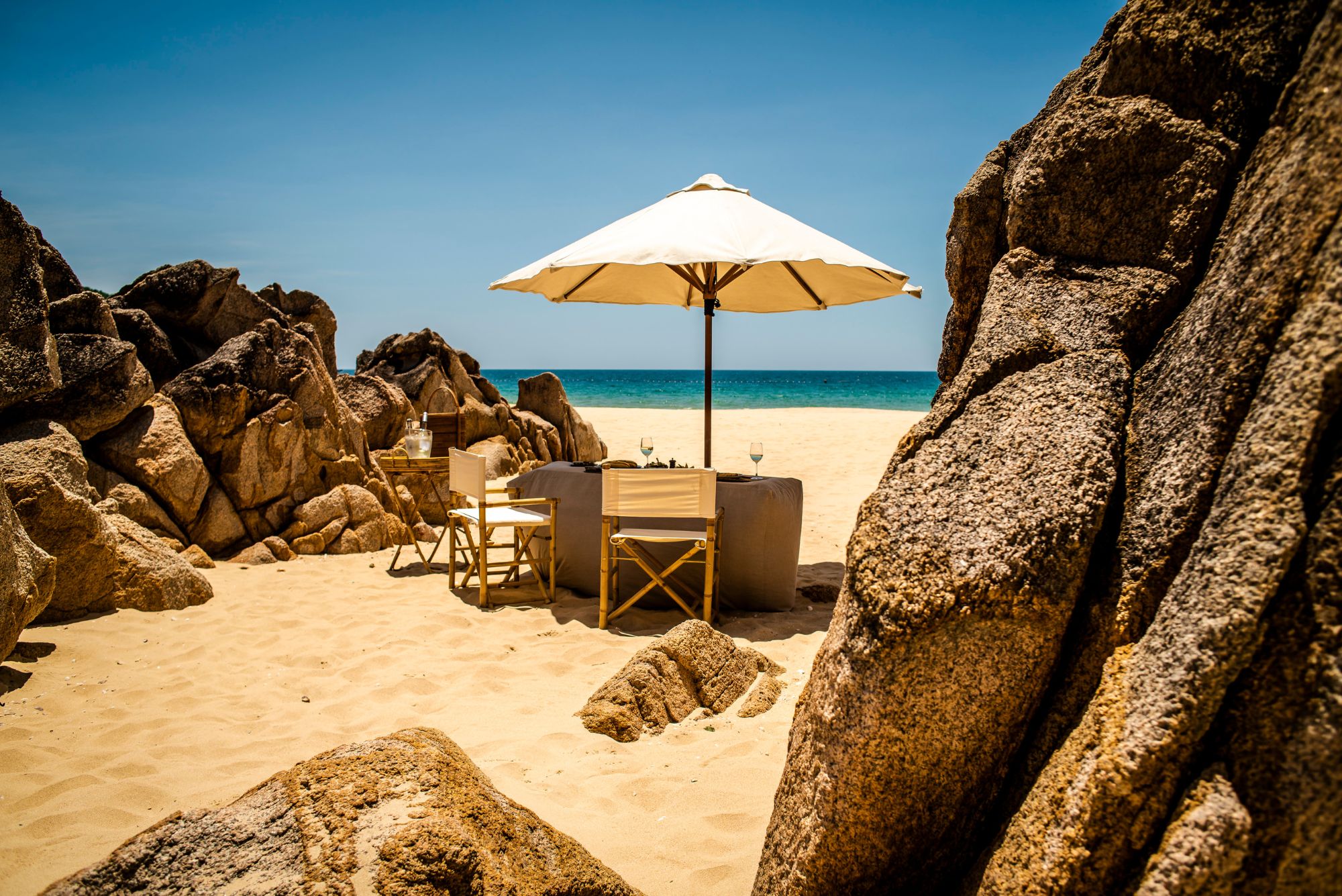An umbrella shades a small table and chairs on Zannier Bãi San Hô's sandy beach, tucked between warm granite rocks.