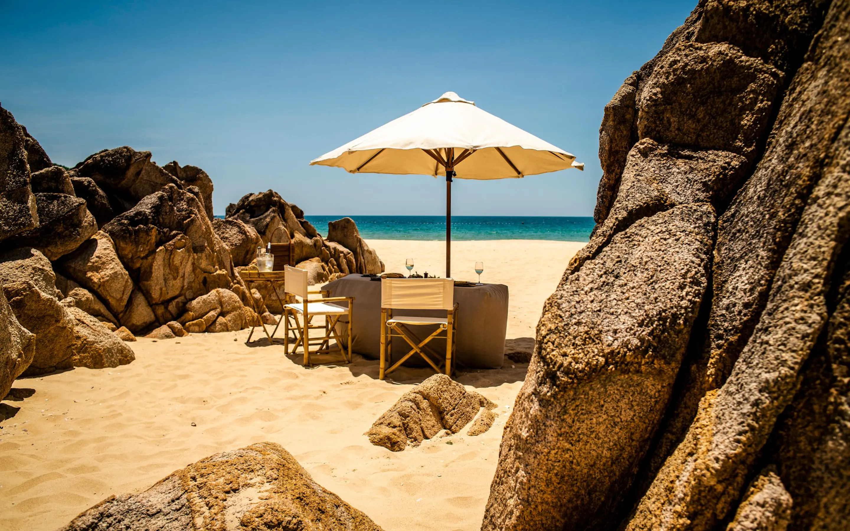 An umbrella shades a small table and chairs on Zannier Bãi San Hô's sandy beach, tucked between warm granite rocks.