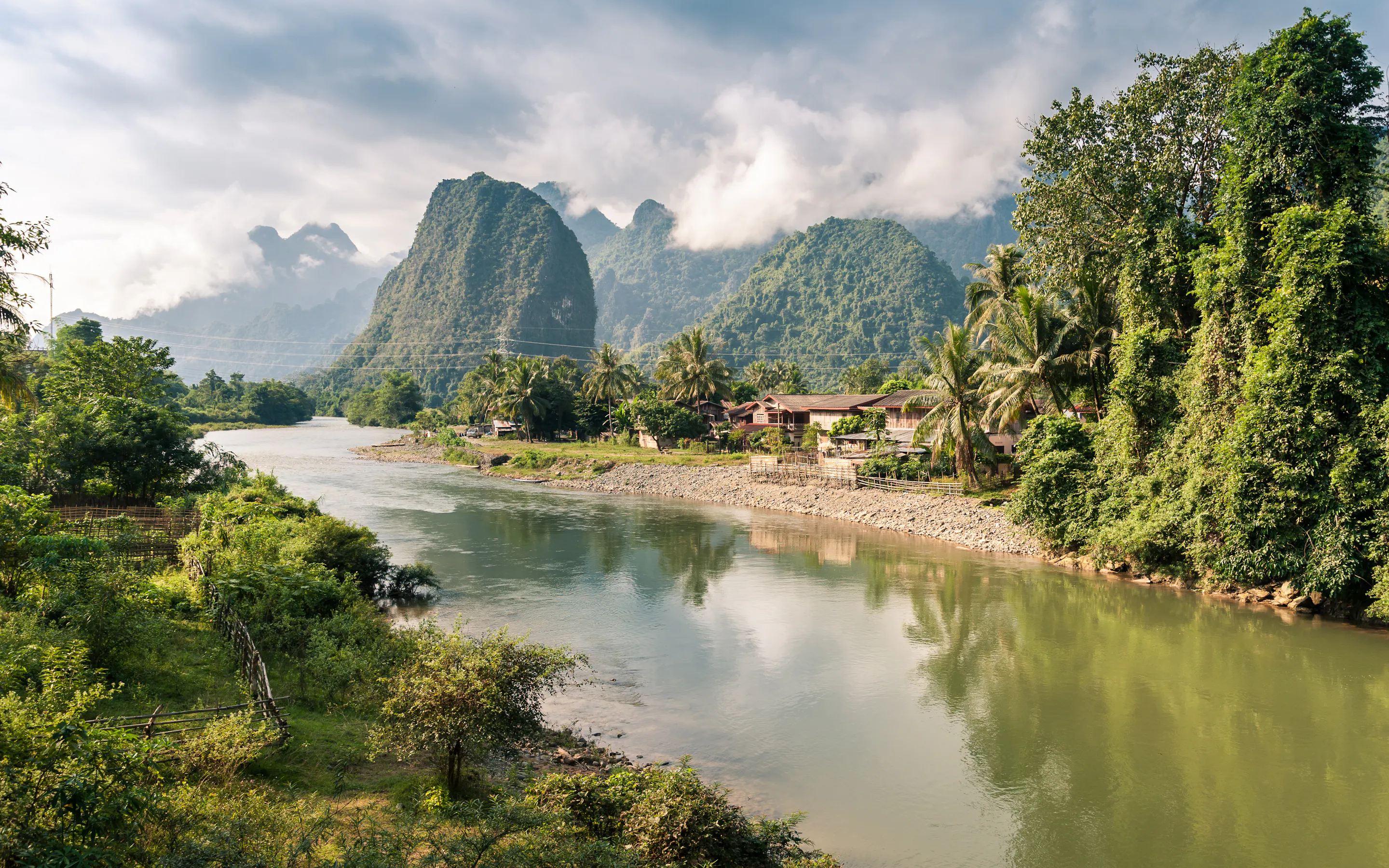 Nam Song River winds past palms and village homes in Vang Vieng, with misty karst mountains rising beyond.