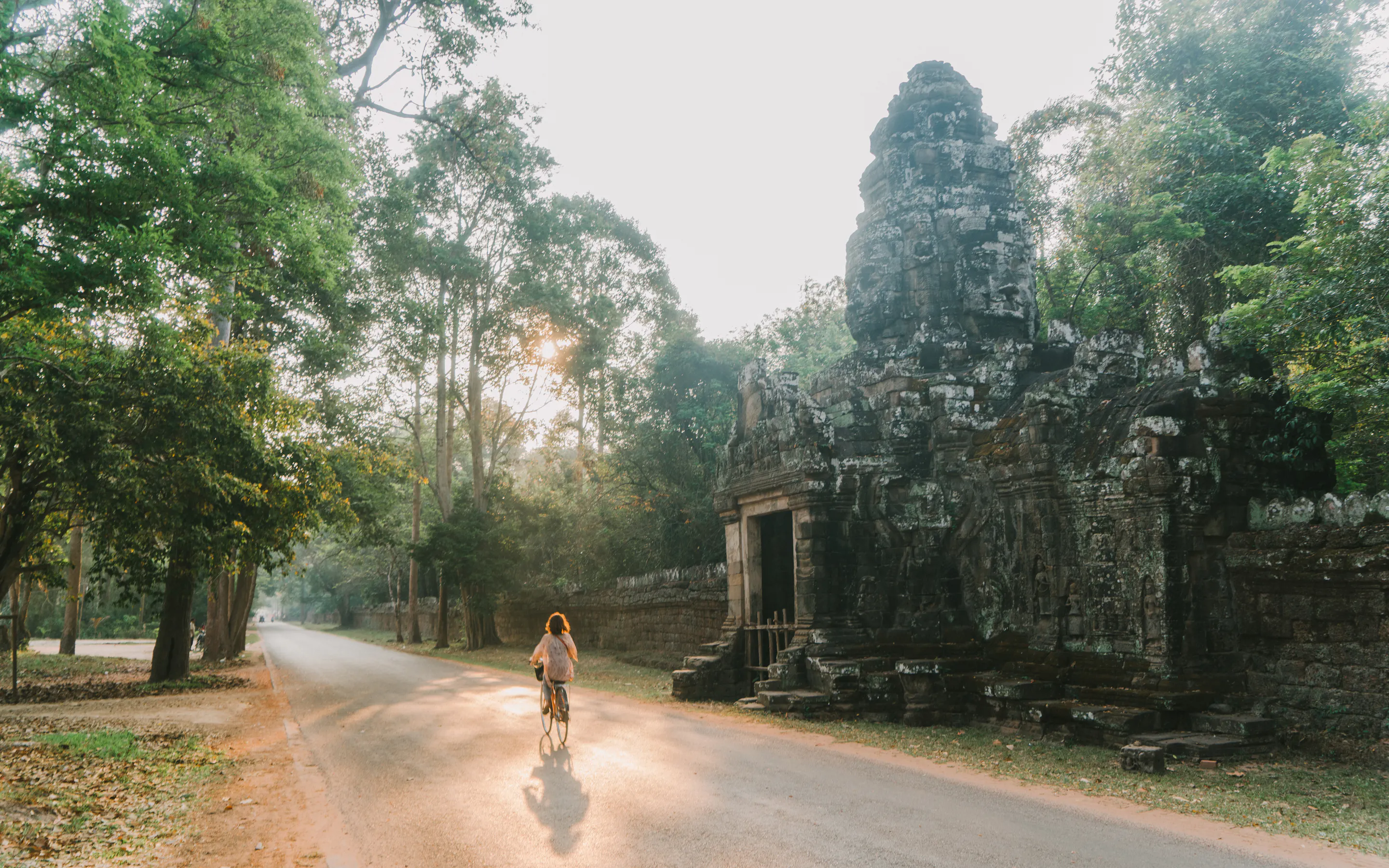 A cyclist rides along a quiet Angkor road beside weathered temple ruins, with sunlight filtering through tall trees.