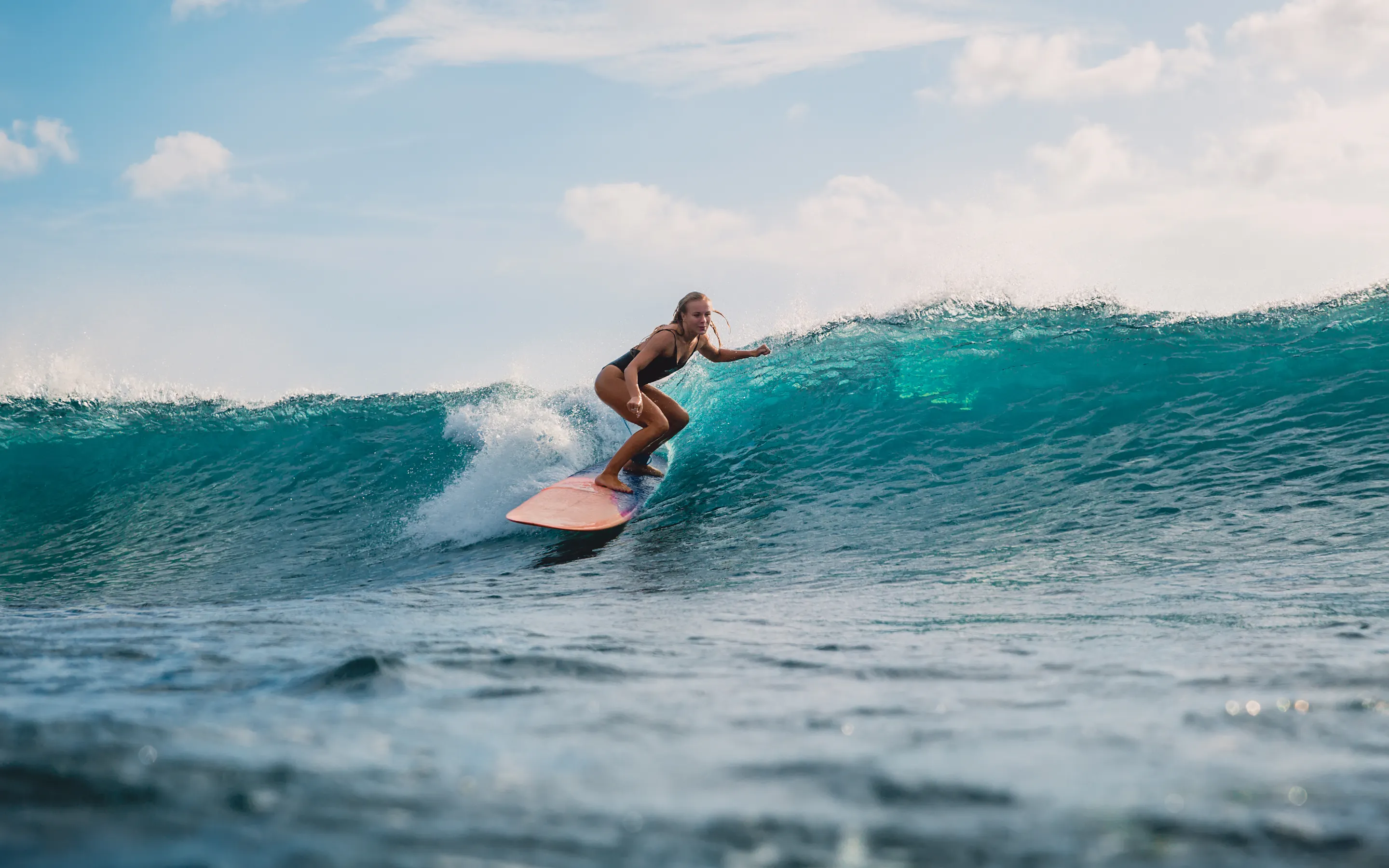 A surfer leans into a bright turquoise wave in Indonesia, with white spray curling across the sunlit water.