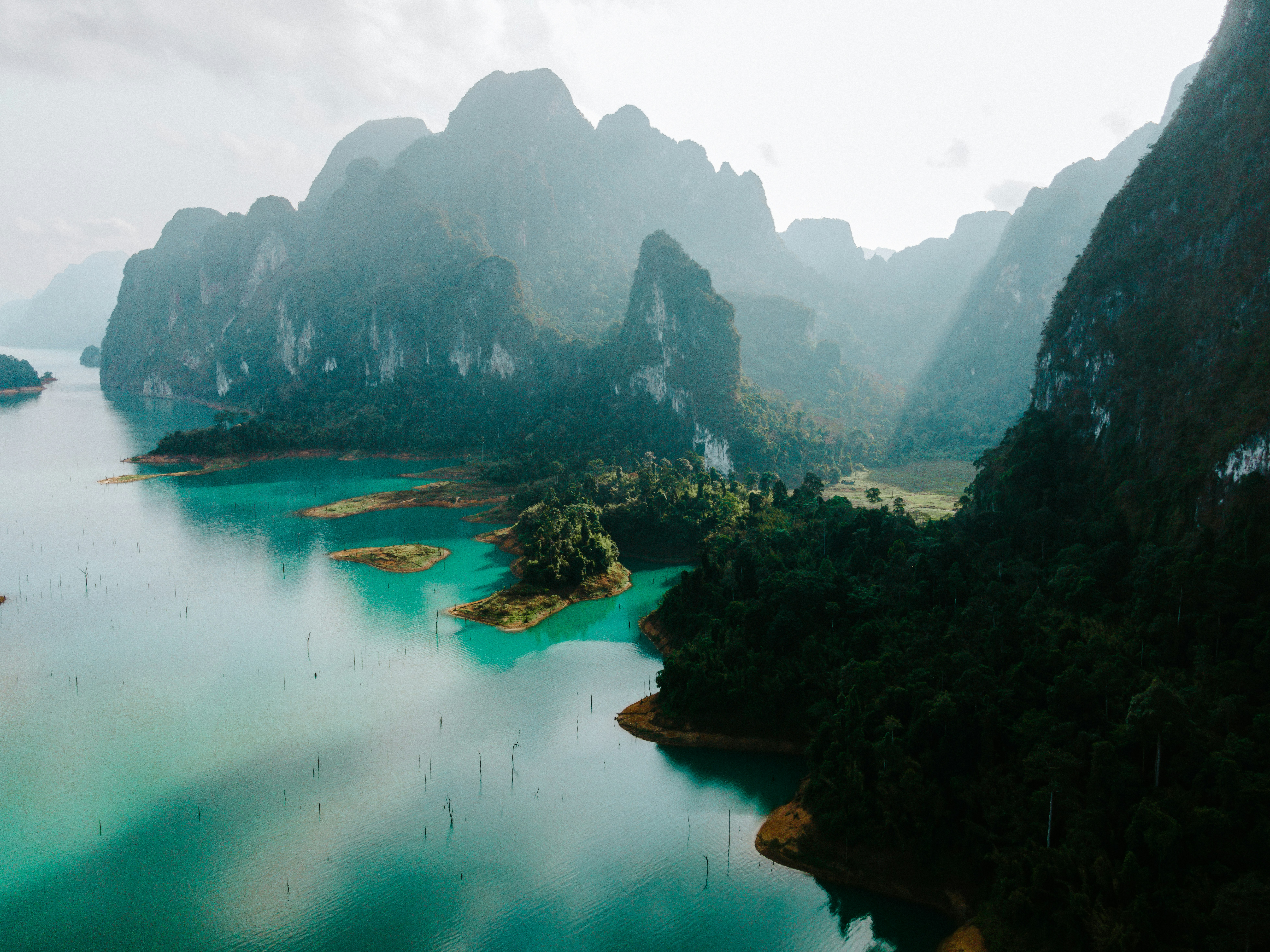 Turquoise water winds through Khao Sok's misty limestone karsts, where forested cliffs rise beneath a pale sky.