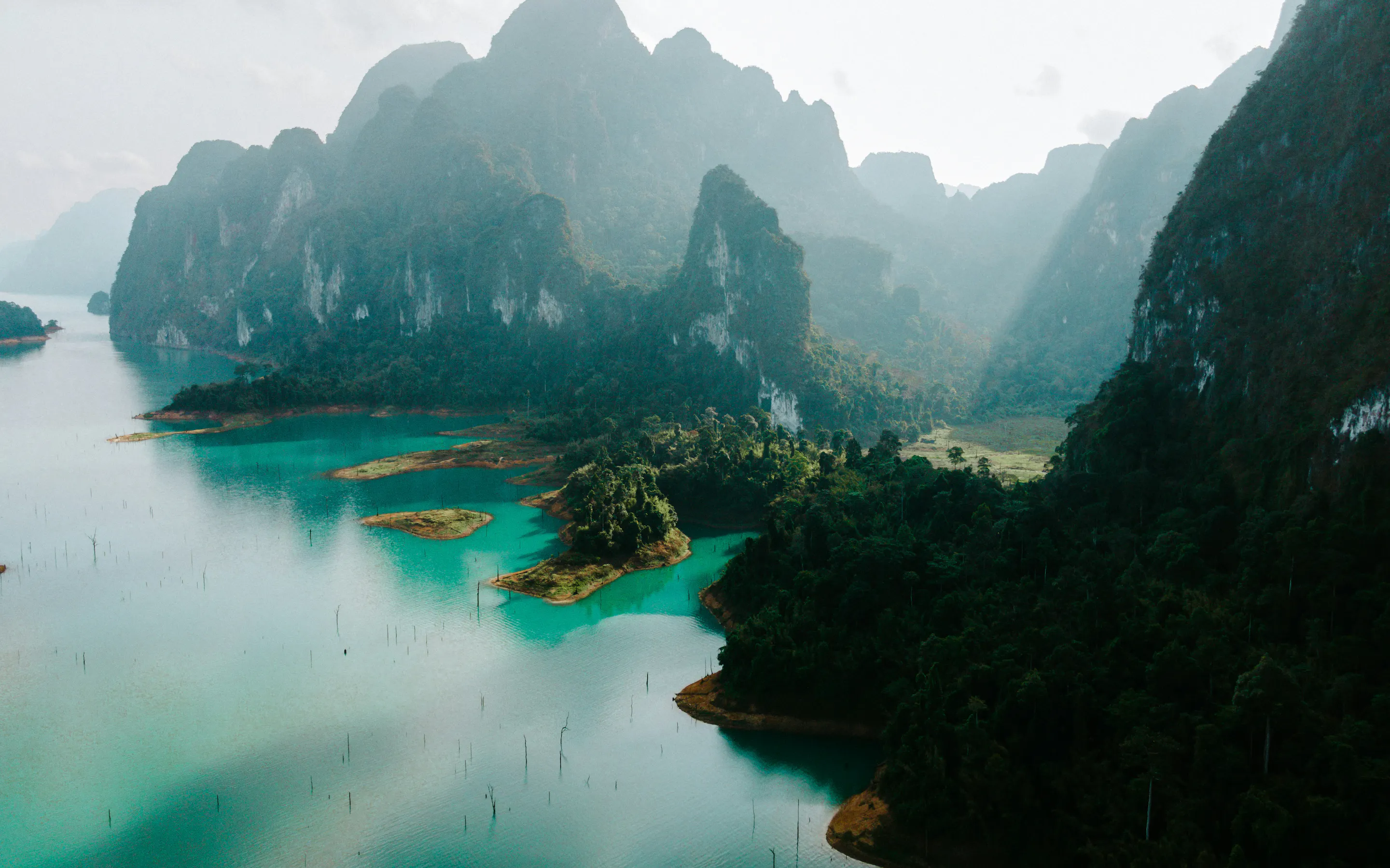 Turquoise water winds through Khao Sok's misty limestone karsts, where forested cliffs rise beneath a pale sky.