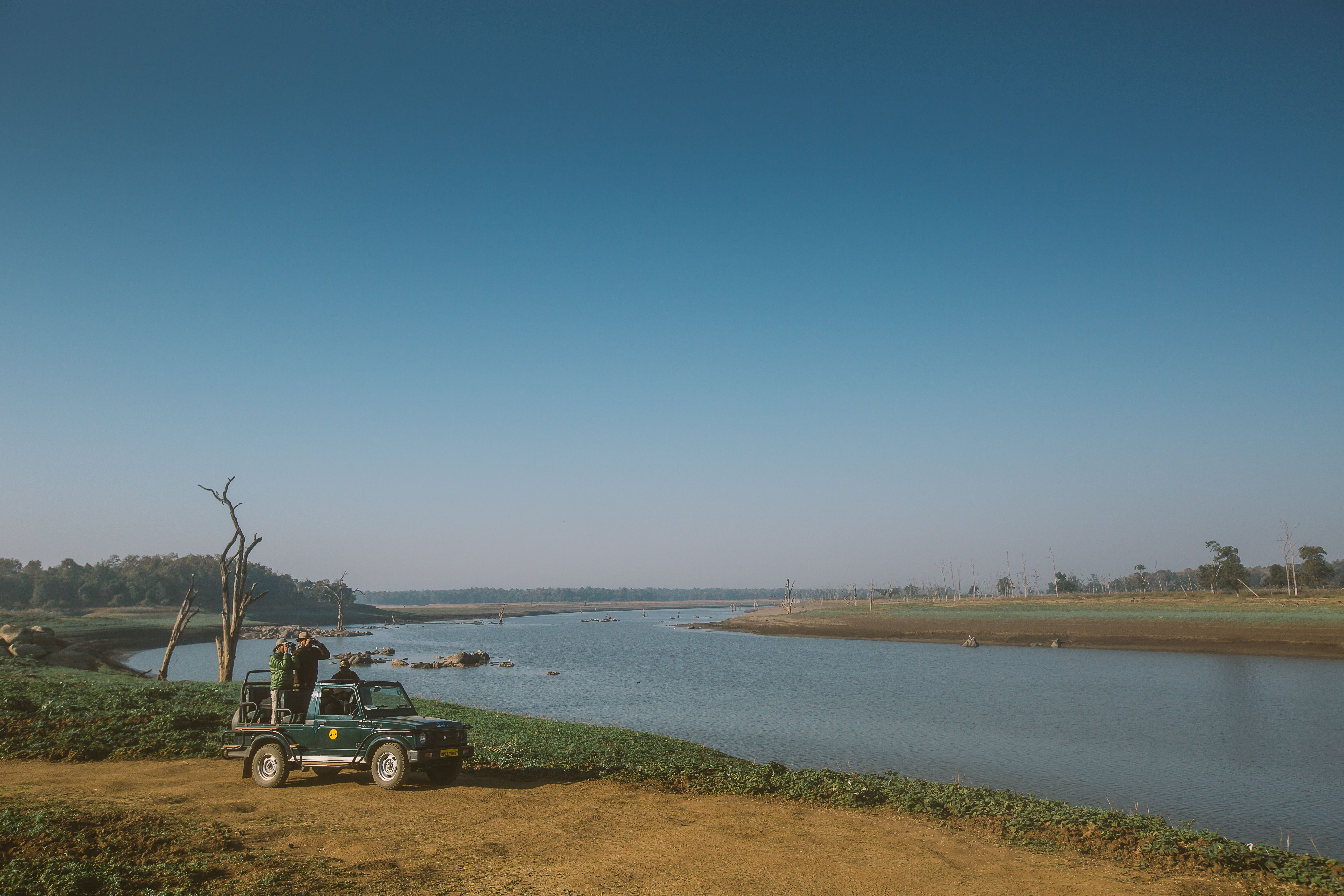 A safari jeep pauses beside a wide river under a clear blue sky, with two guests standing up to scan the banks.