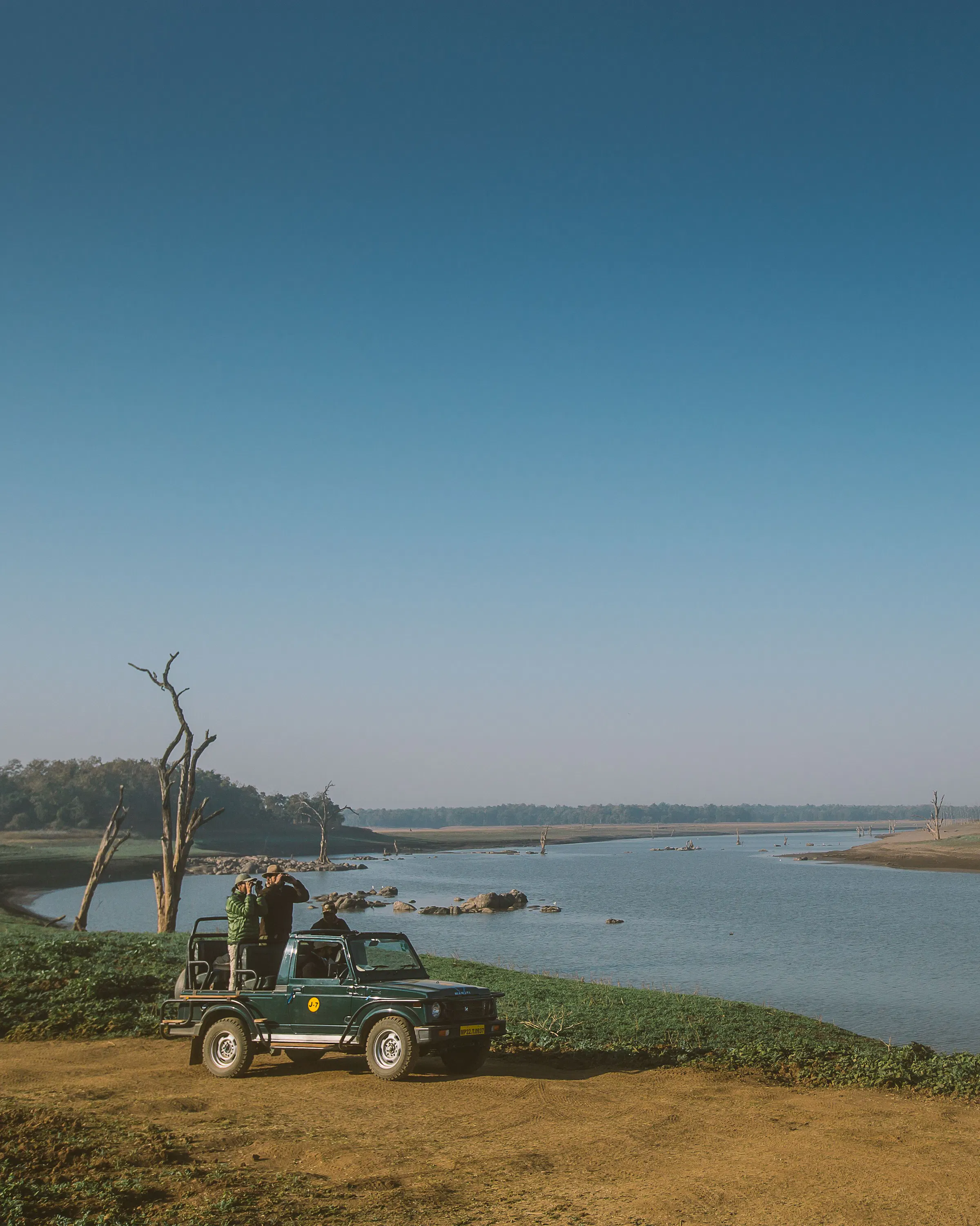 A safari jeep pauses beside a wide river under a clear blue sky, with two guests standing up to scan the banks.