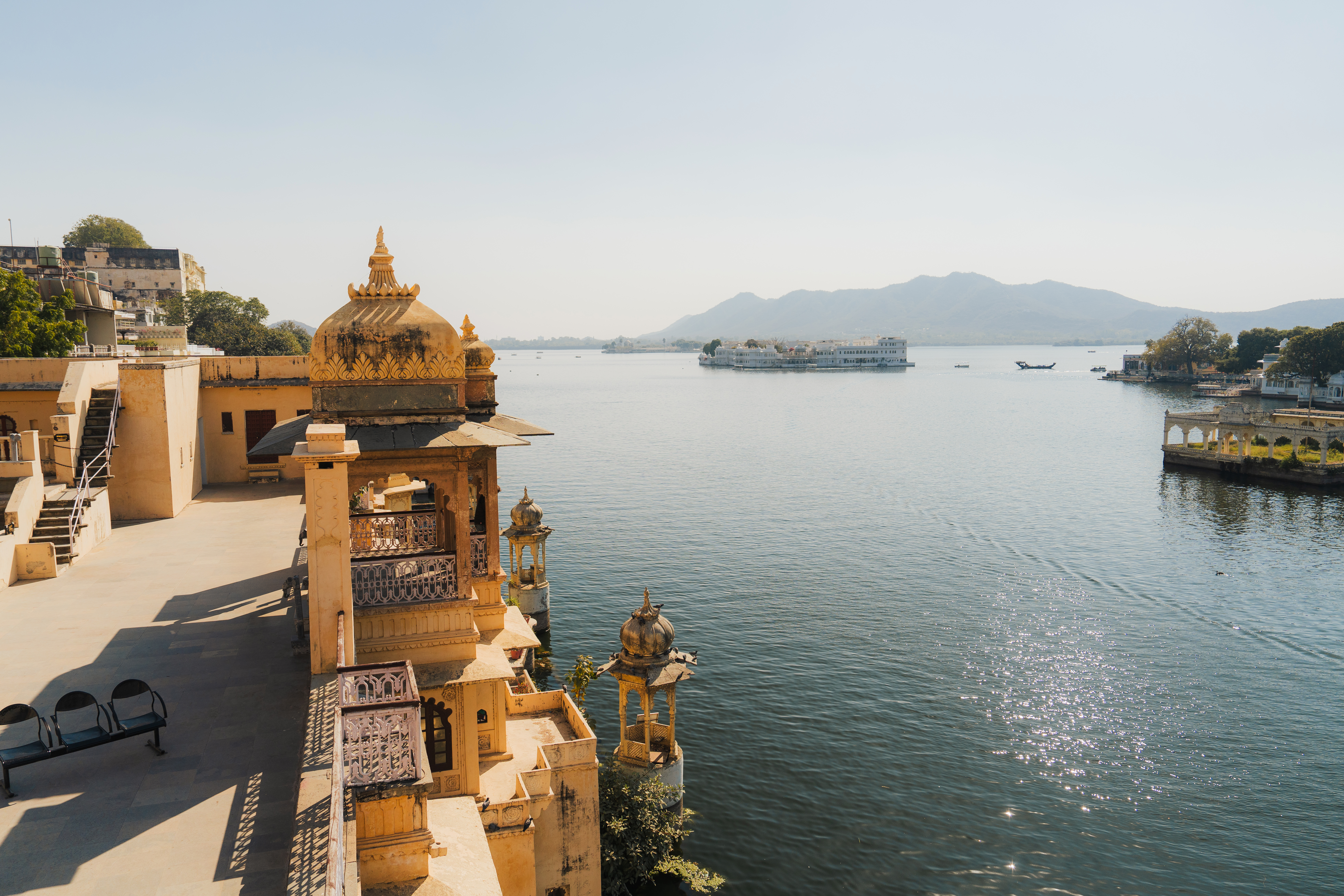 Golden rooftops and pale stone pavilions overlook Lake Pichola at sunset, with still water, boats, and hazy hills.
