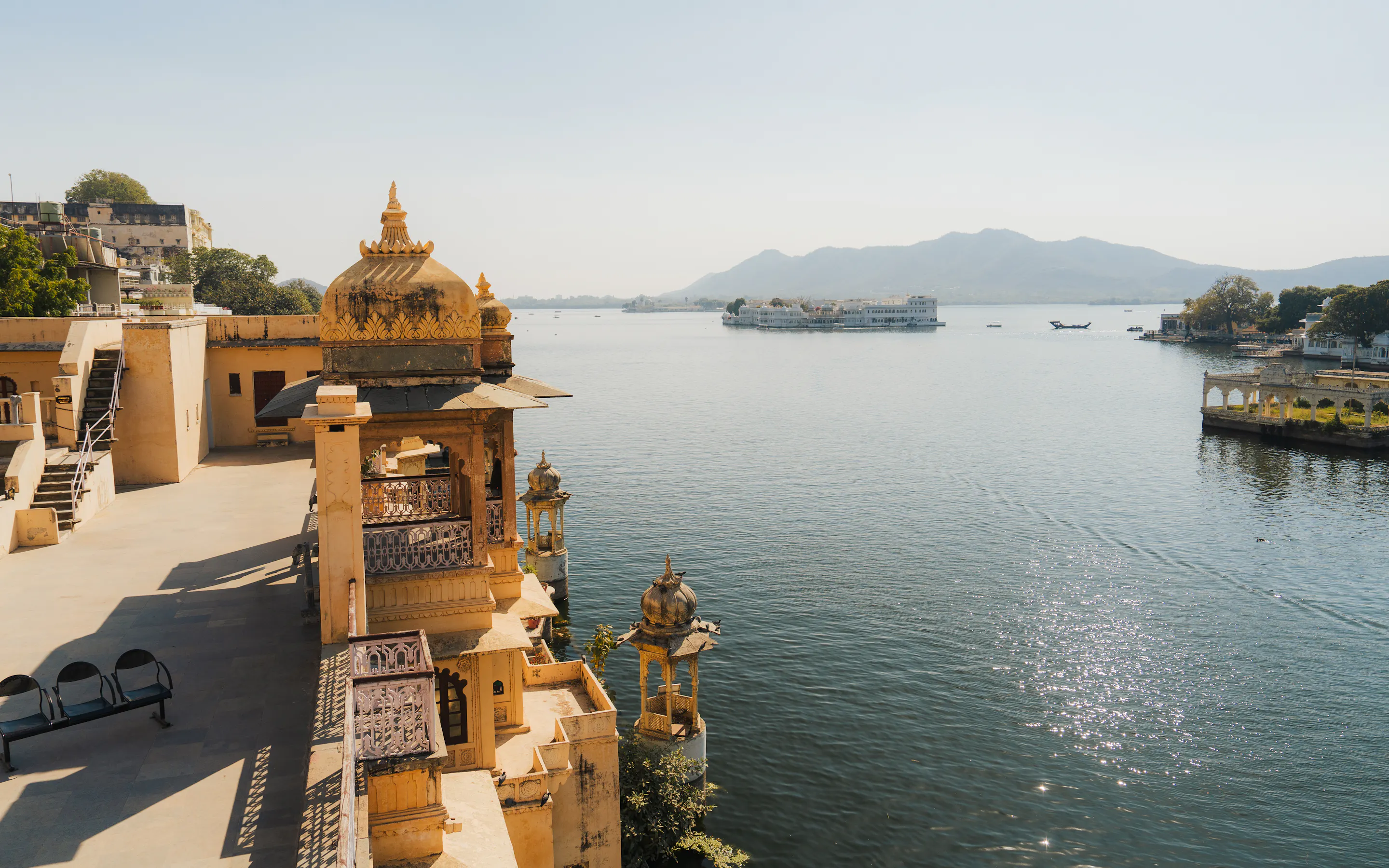 Golden rooftops and pale stone pavilions overlook Lake Pichola at sunset, with still water, boats, and hazy hills.