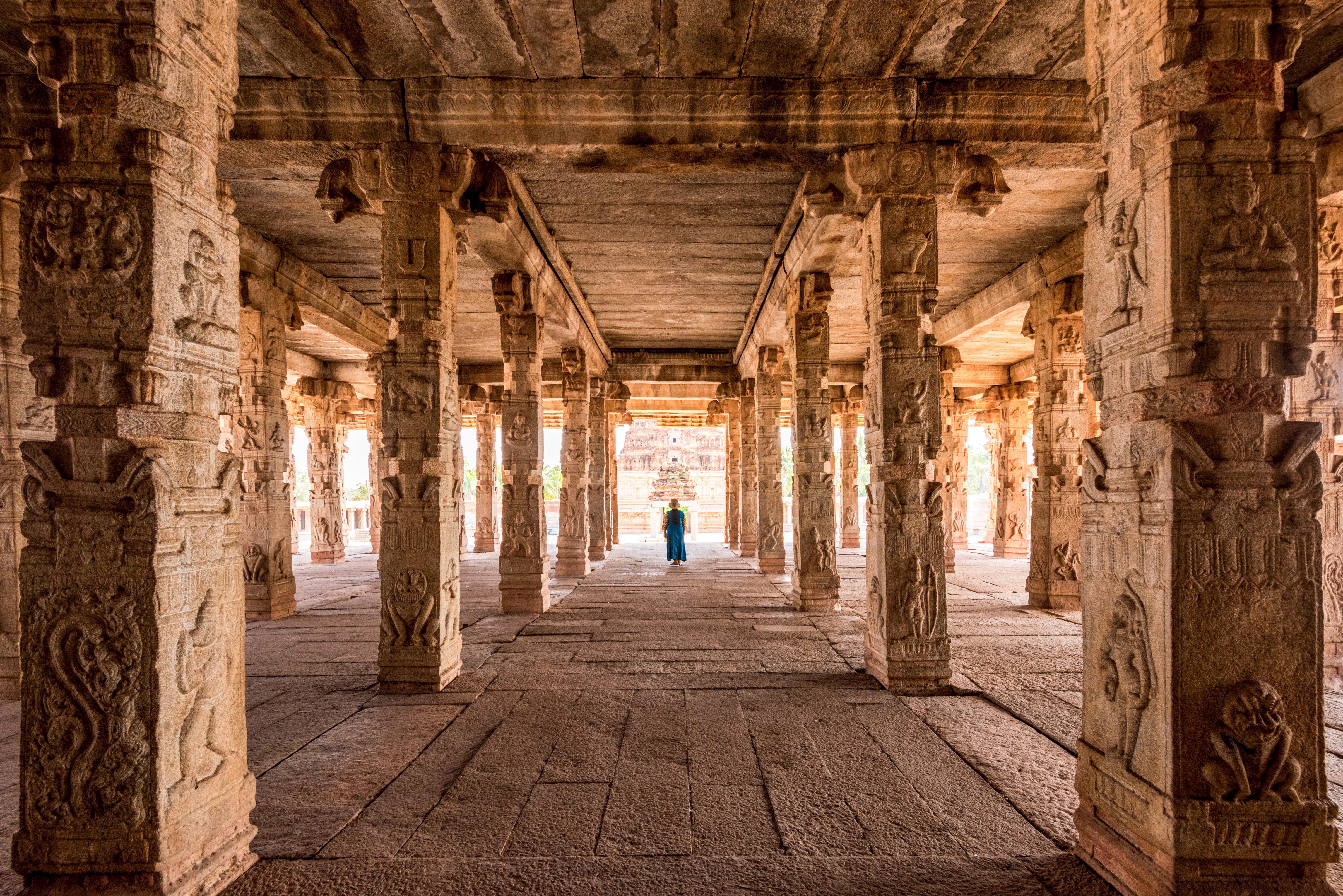 Carved stone columns frame a long temple hall in Hampi, where one visitor stands beneath warm light at the far end.