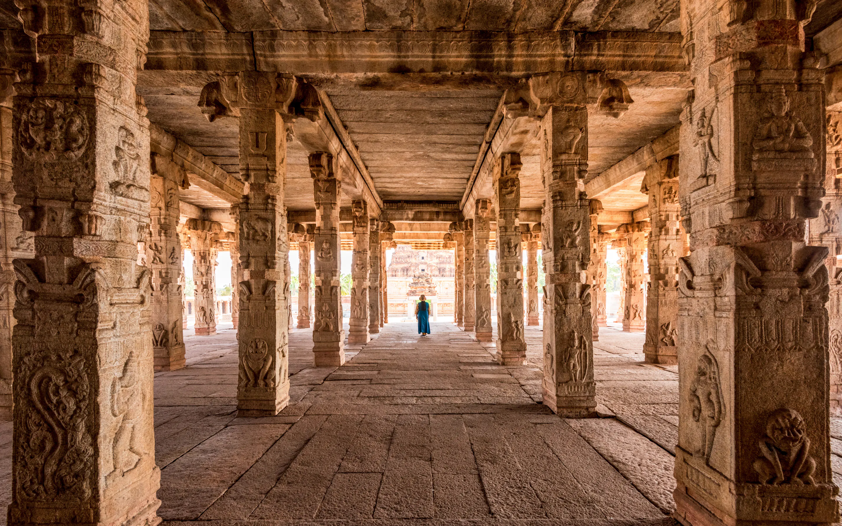 Carved stone columns frame a long temple hall in Hampi, where one visitor stands beneath warm light at the far end.
