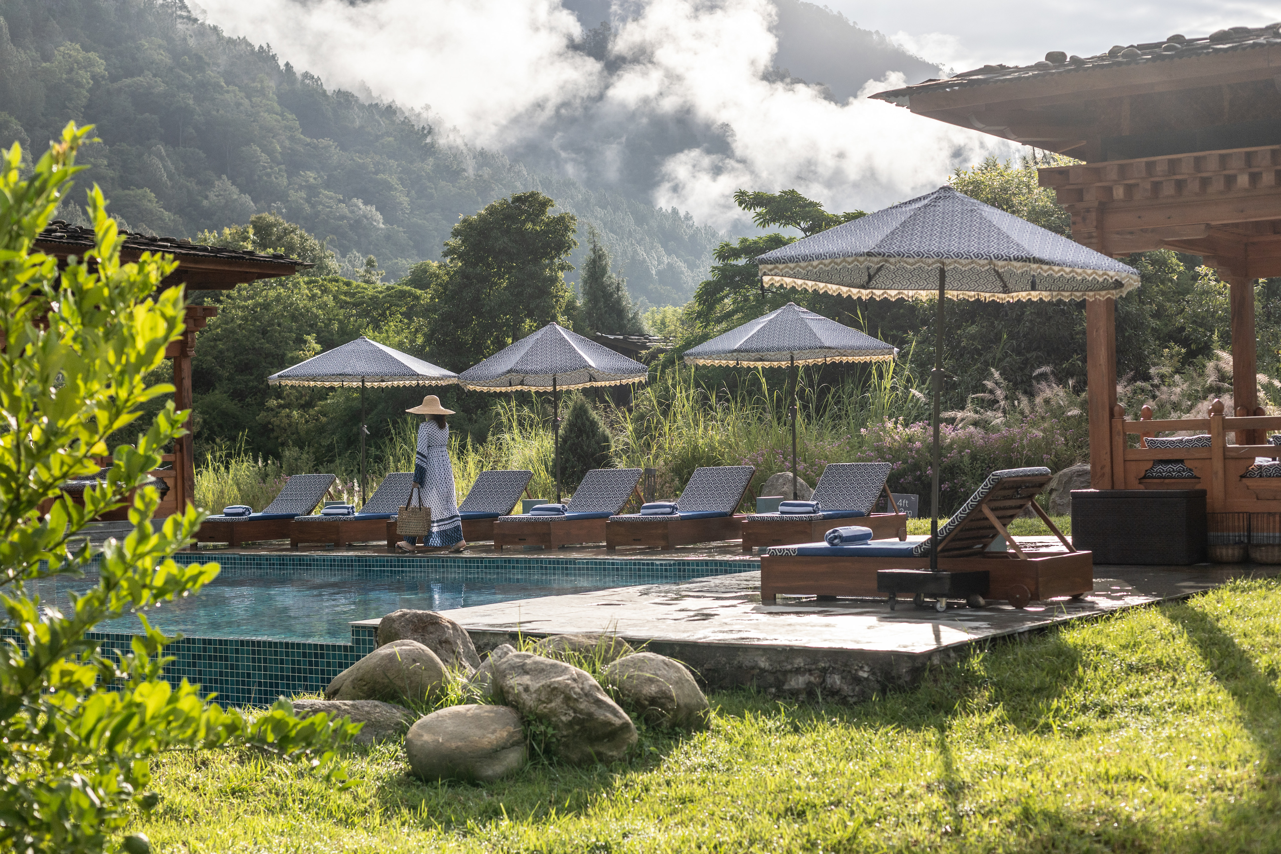 A riverside pool terrace at Punakha River Lodge sits among forested hills, with loungers, umbrellas, and warm sun.
