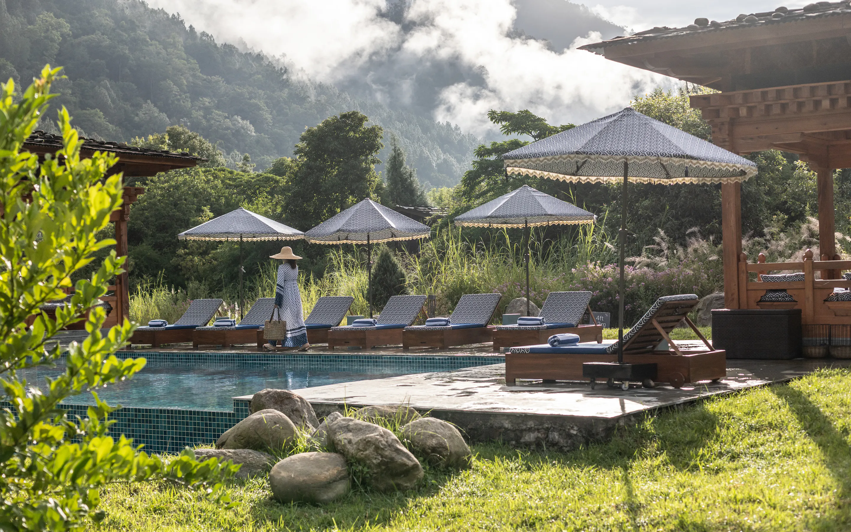 A riverside pool terrace at Punakha River Lodge sits among forested hills, with loungers, umbrellas, and warm sun.
