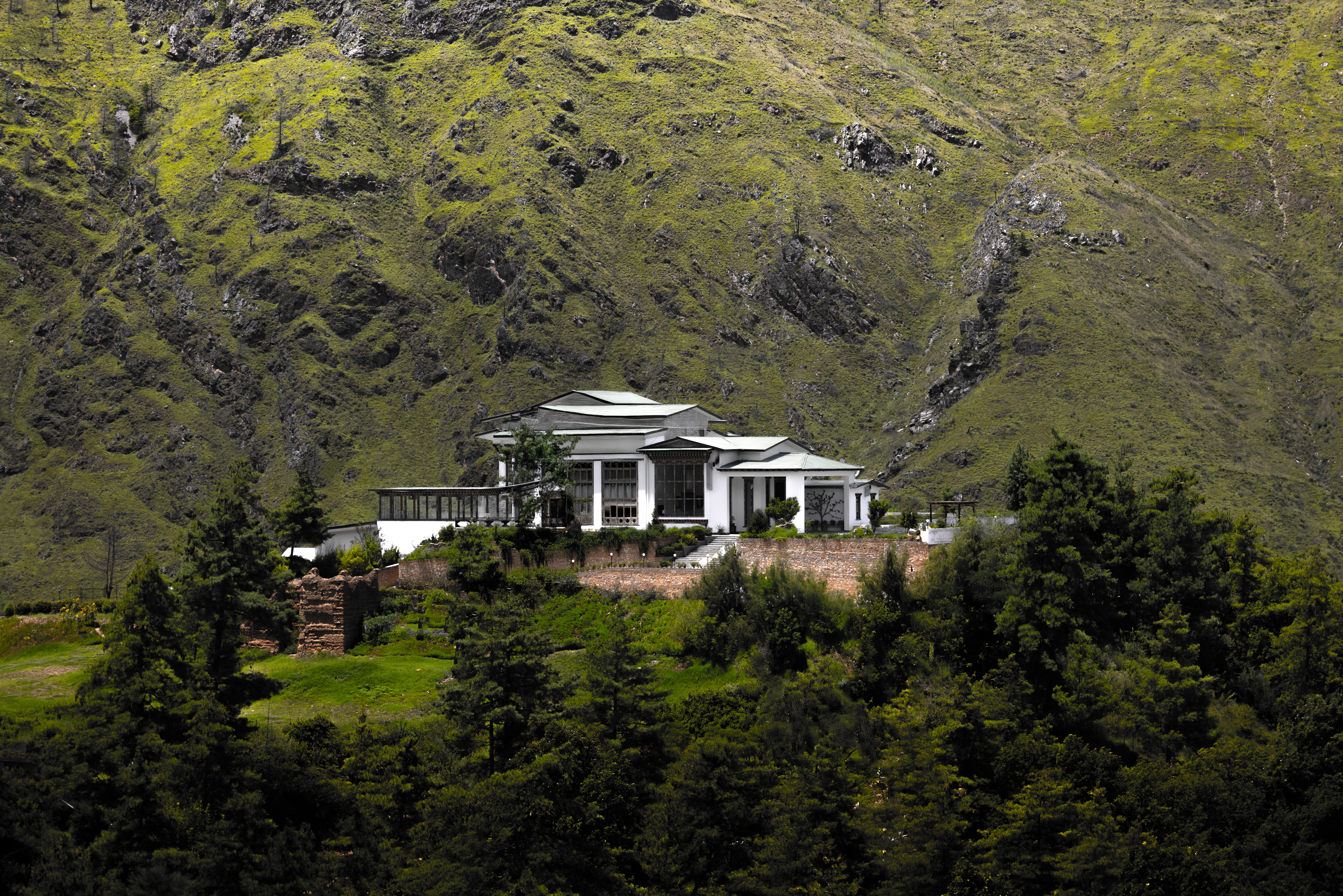 The Postcard Dewa stands above tall trees on a steep green hillside, with rocky mountain slopes rising behind.