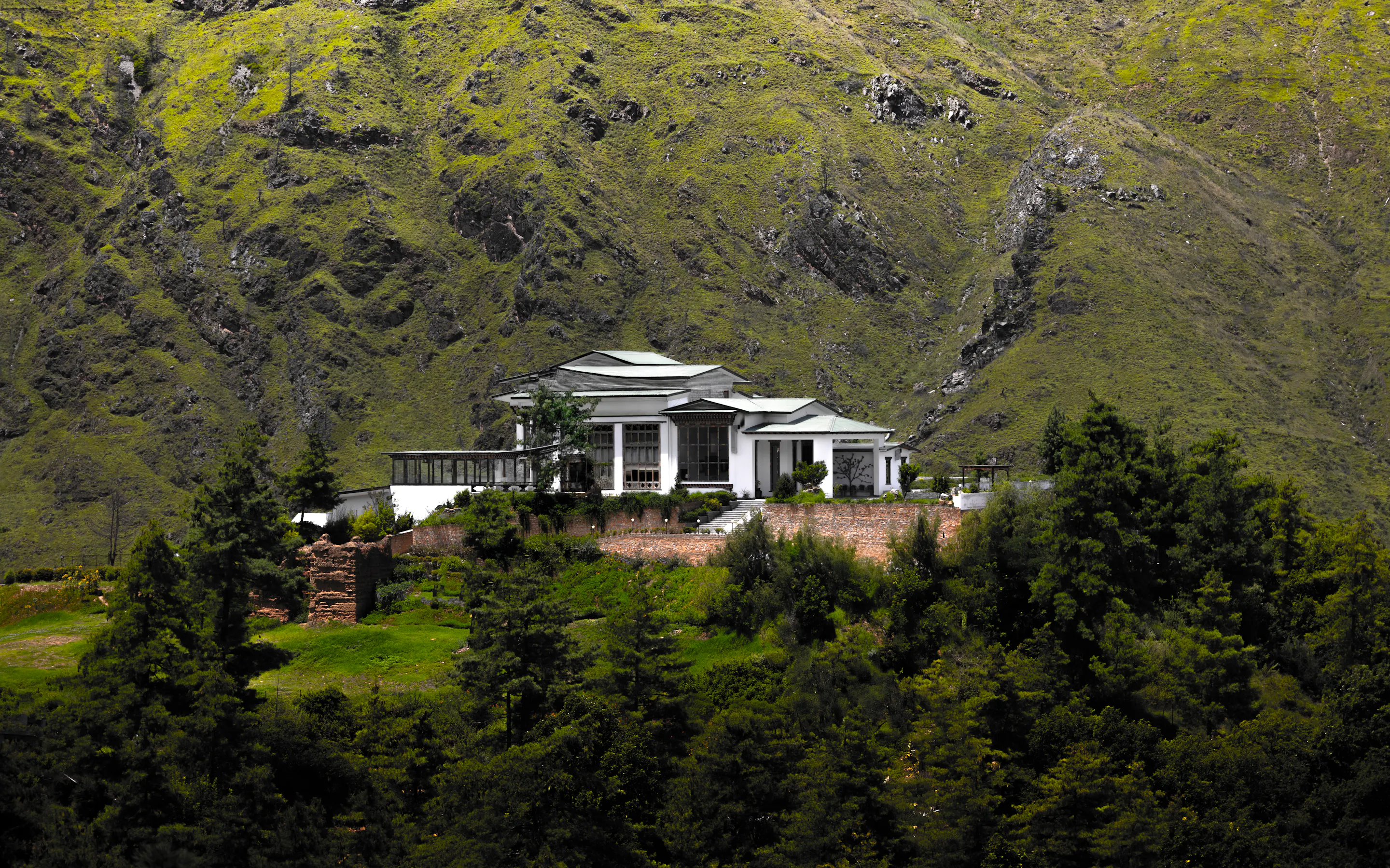The Postcard Dewa stands above tall trees on a steep green hillside, with rocky mountain slopes rising behind.