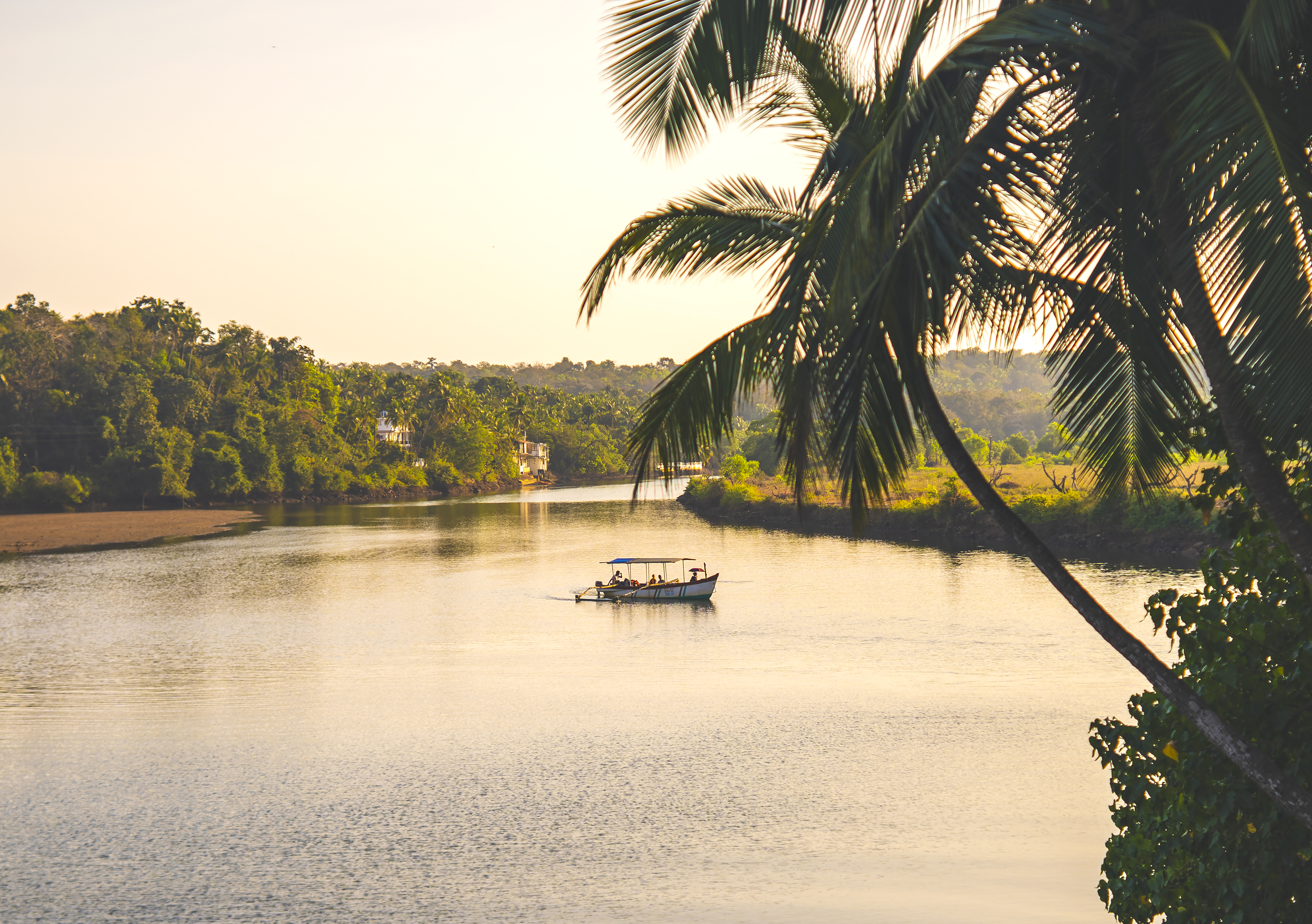 A small boat drifts along a calm South Goa river at sunset, framed by palms and still water in warm evening light.