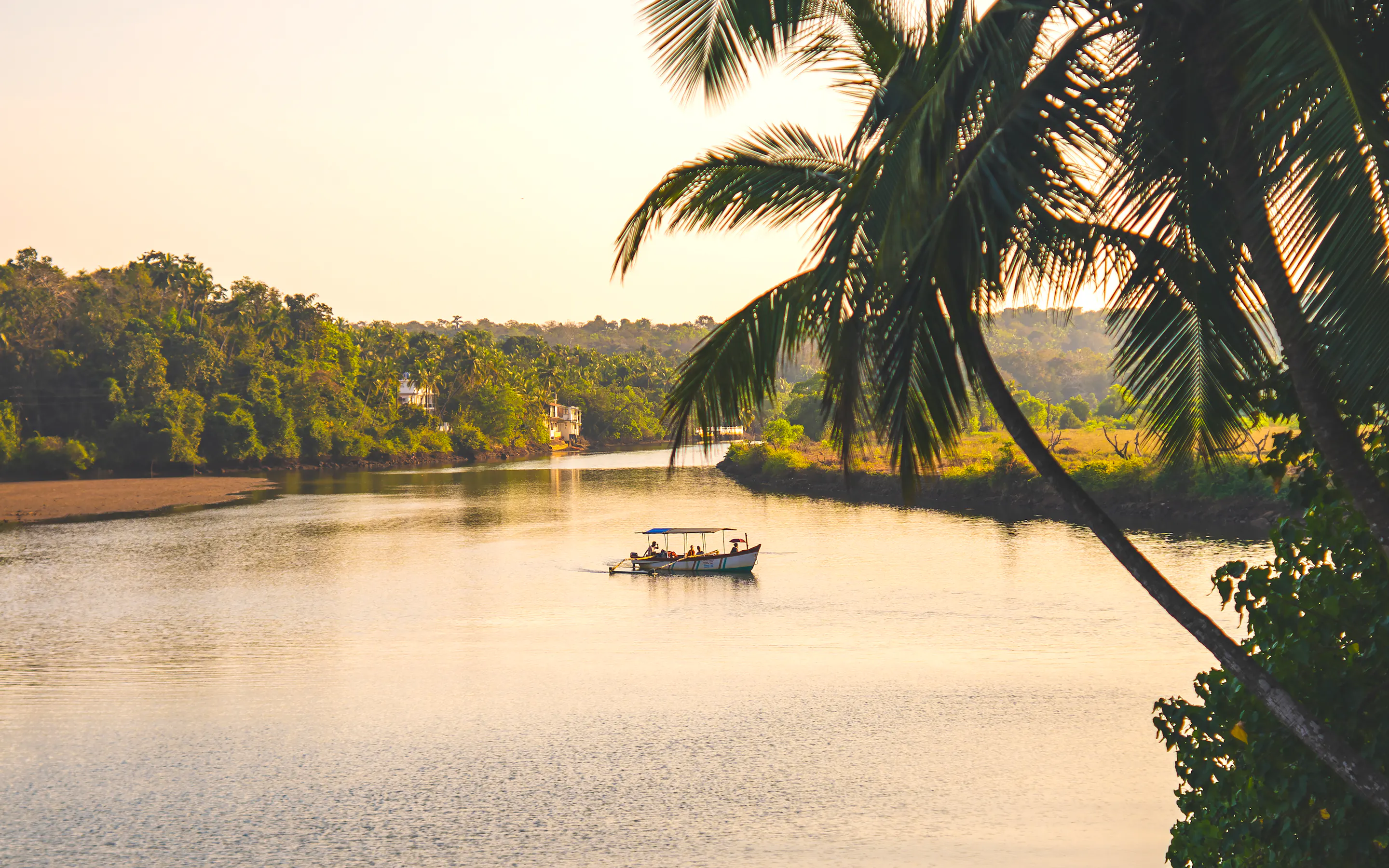 A small boat drifts along a calm South Goa river at sunset, framed by palms and still water in warm evening light.
