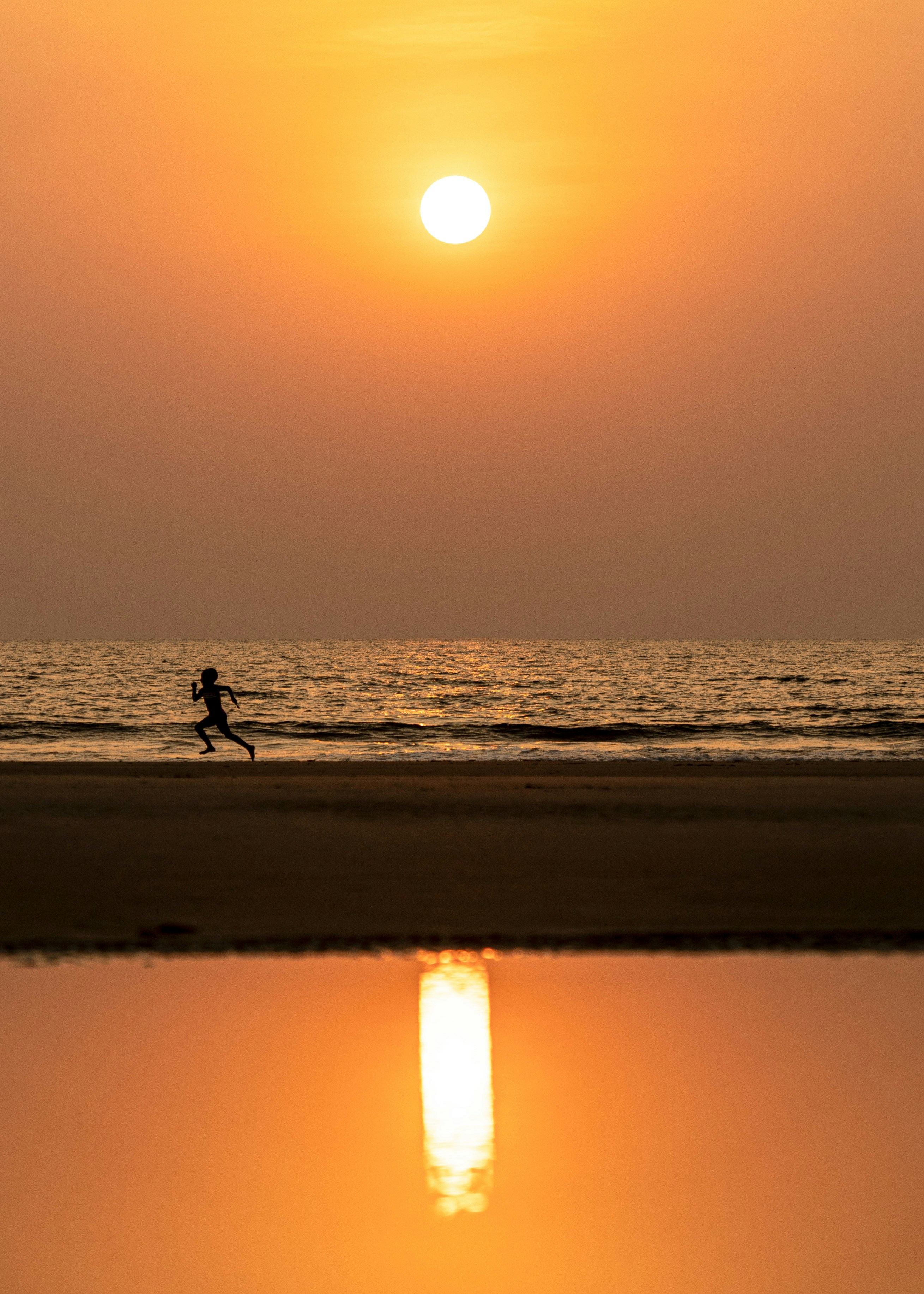 One person runs along the shoreline at sunset, with the low sun mirrored in wet sand beneath a hazy orange sky.