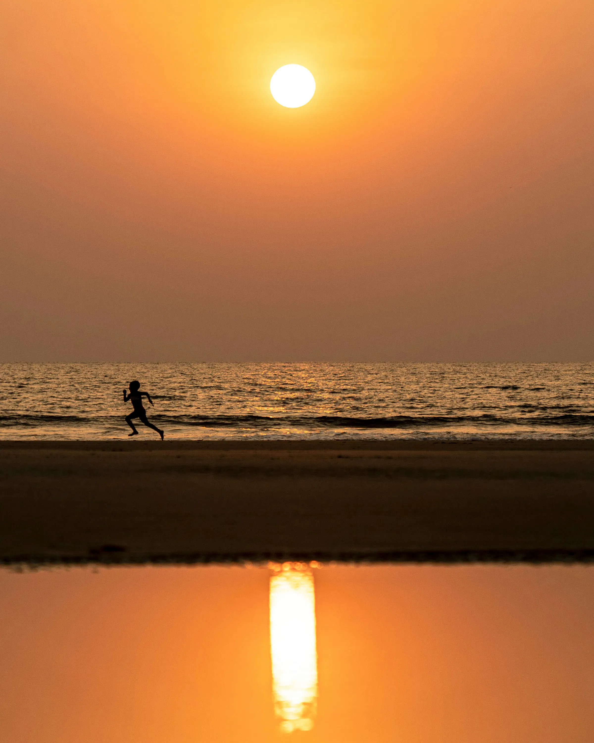 One person runs along the shoreline at sunset, with the low sun mirrored in wet sand beneath a hazy orange sky.