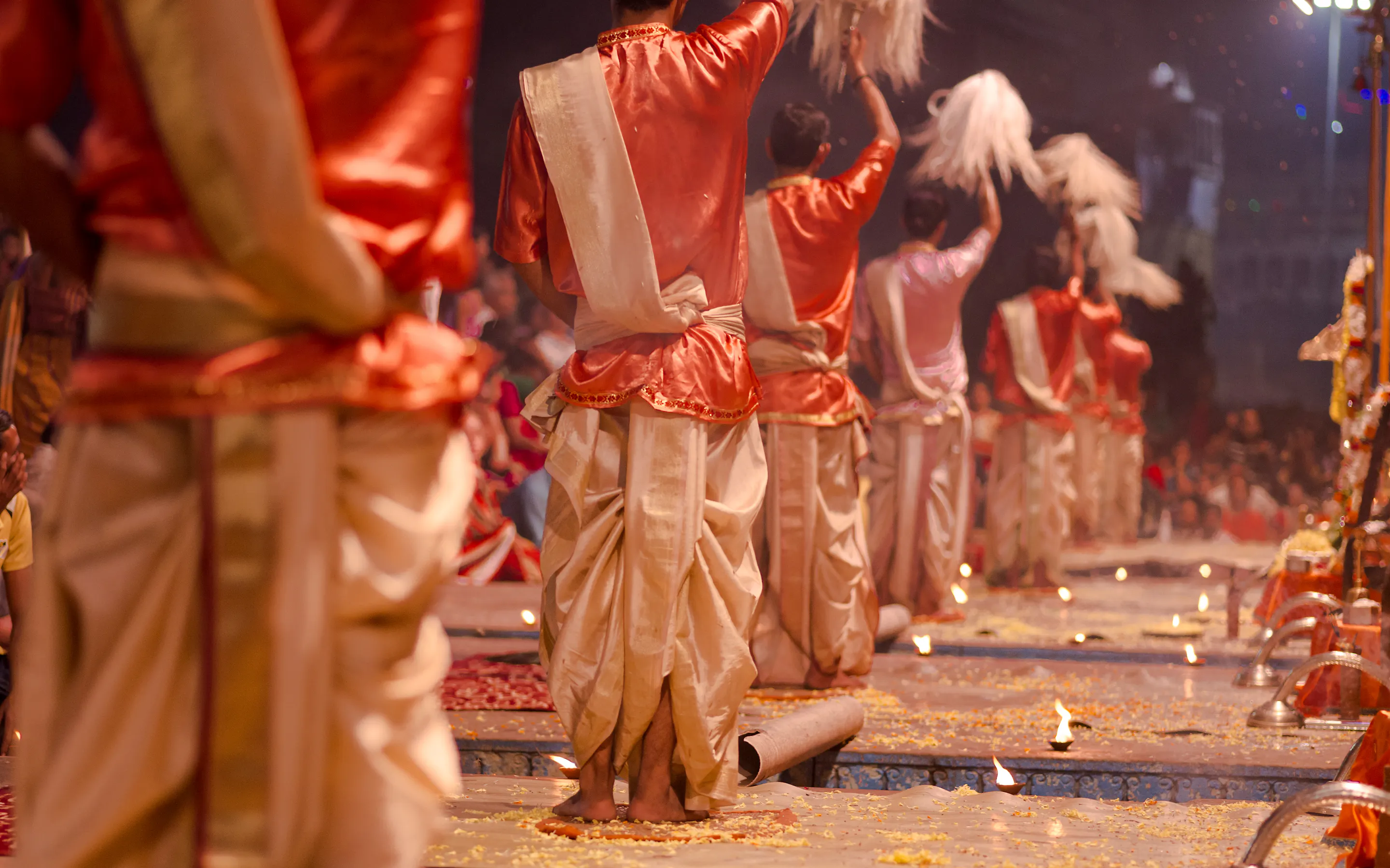 Priests perform an evening puja ceremony in Varanasi, with raised fly whisks, warm light, and rows of small flames.