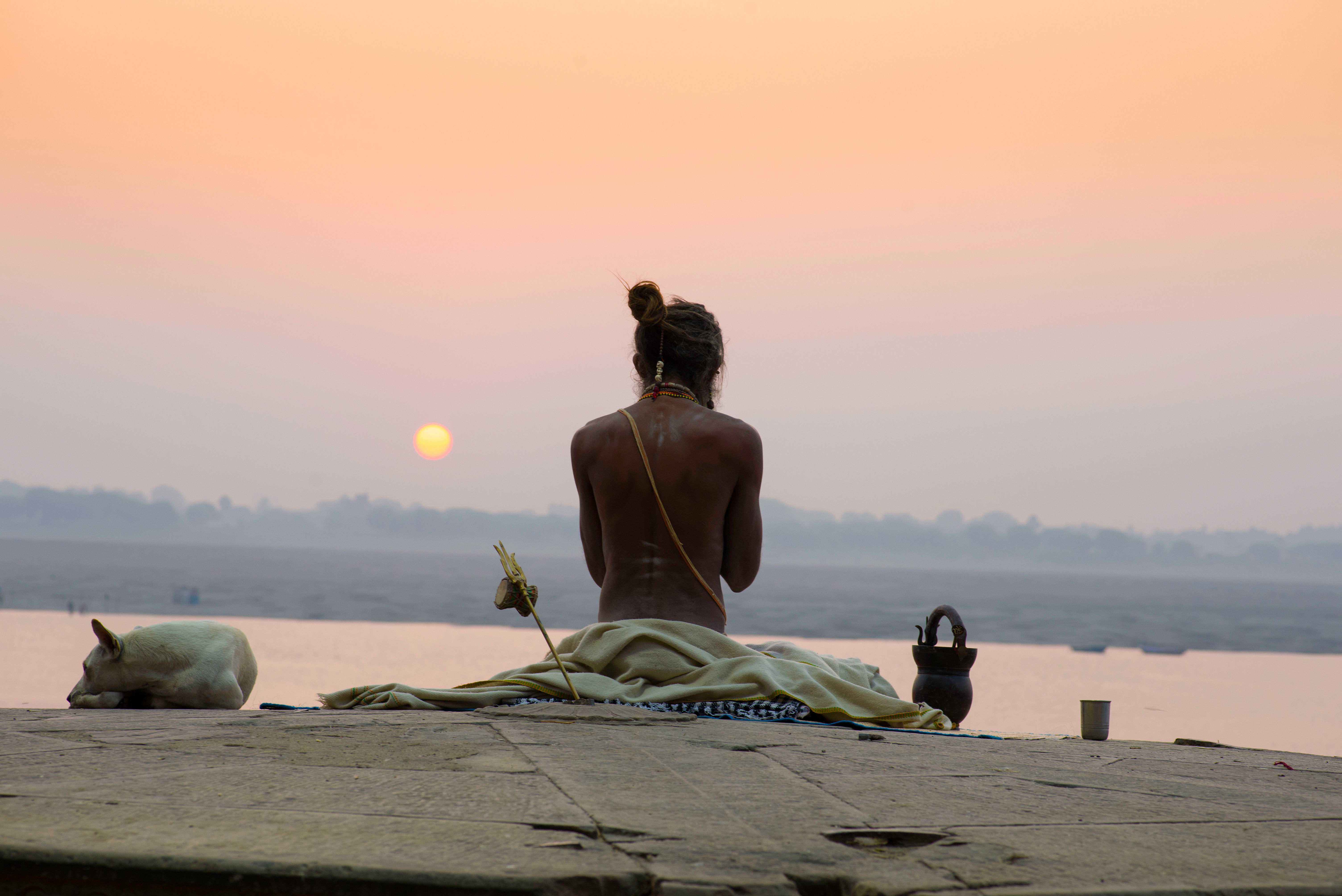 A man sits on the Varanasi riverfront at sunrise beside a resting dog, with calm water, boats, and a pale orange sky.