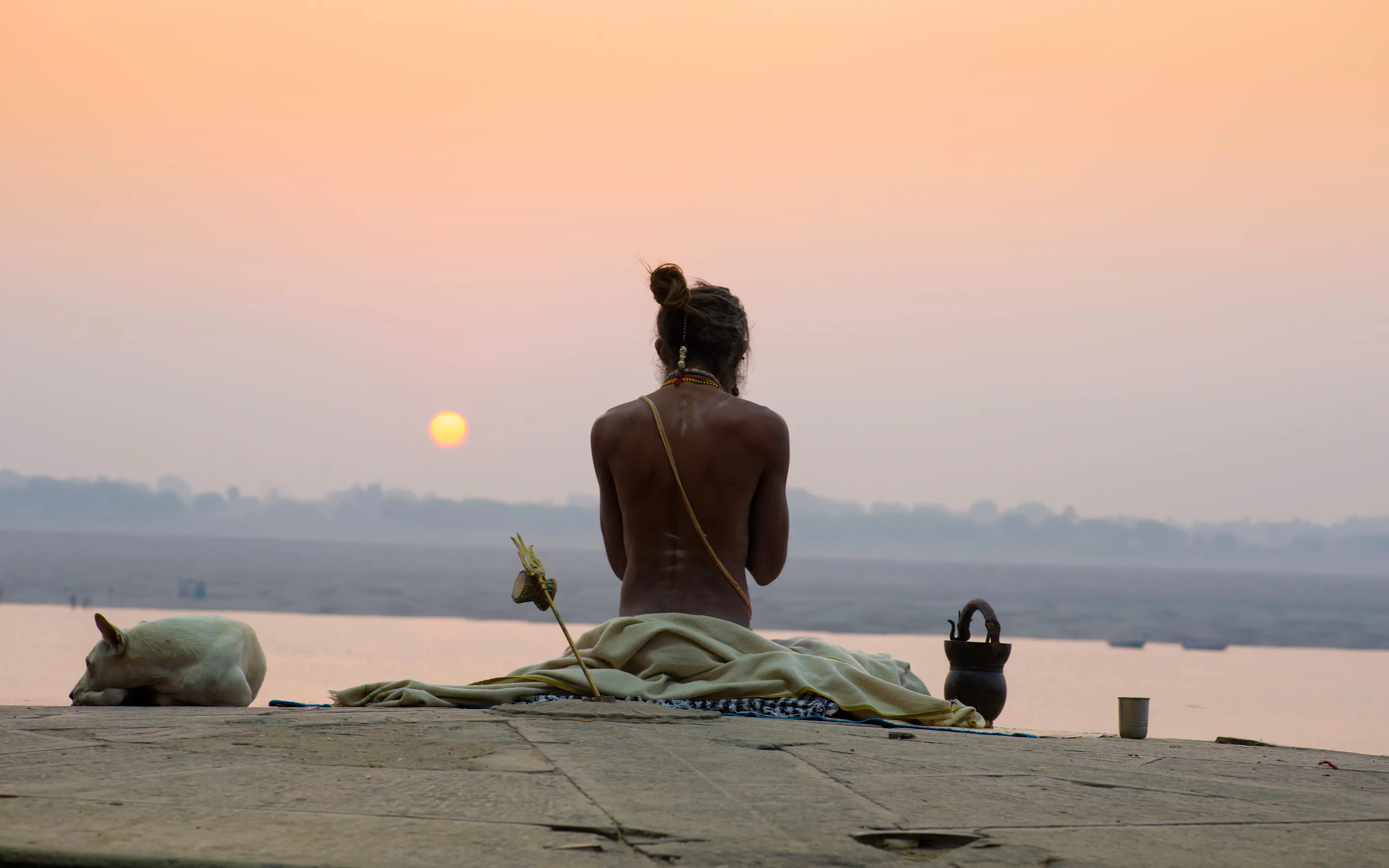 A man sits on the Varanasi riverfront at sunrise beside a resting dog, with calm water, boats, and a pale orange sky.