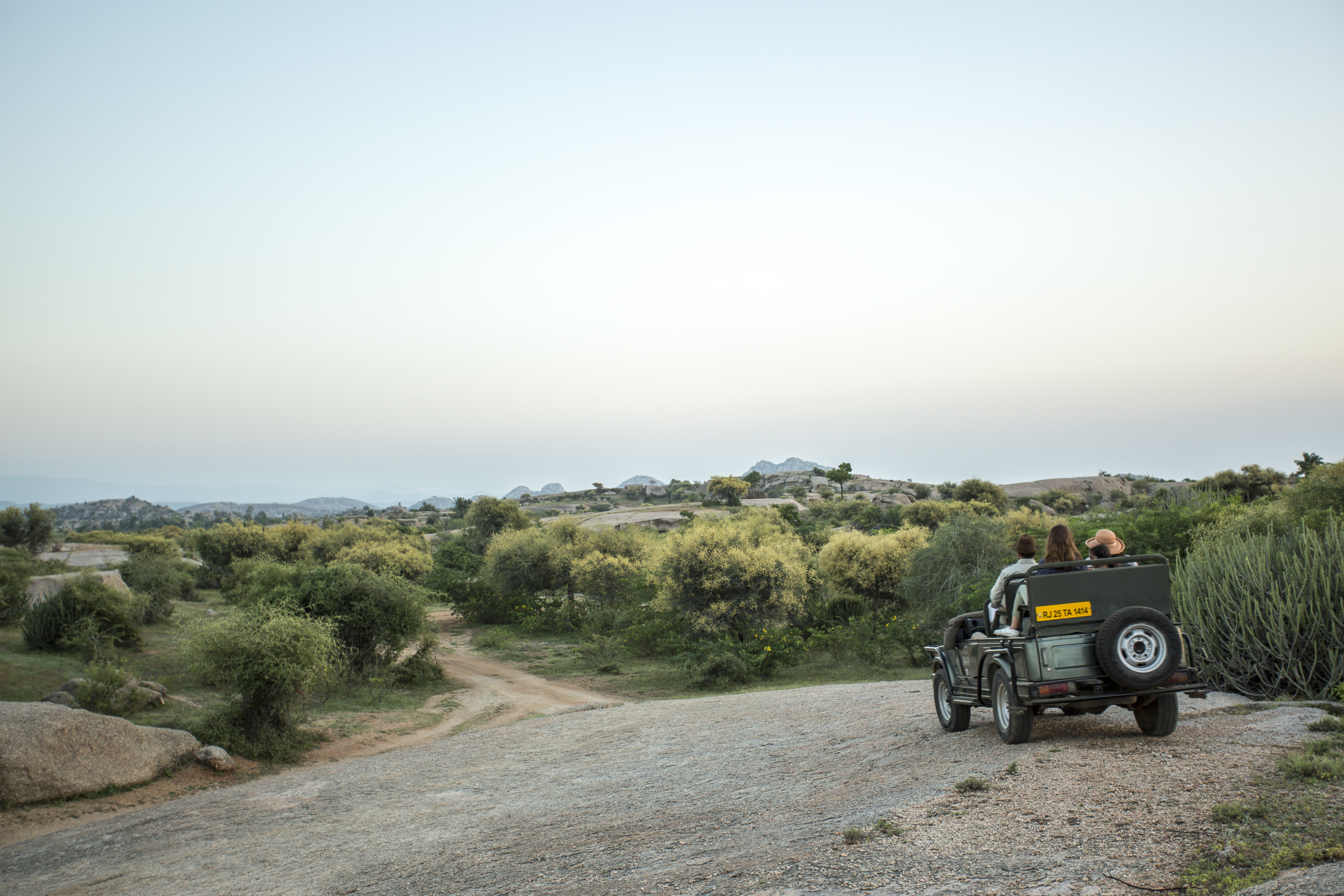 A safari jeep pauses on a gravel track above low scrub, with two guests seated inside and rounded hills beyond.