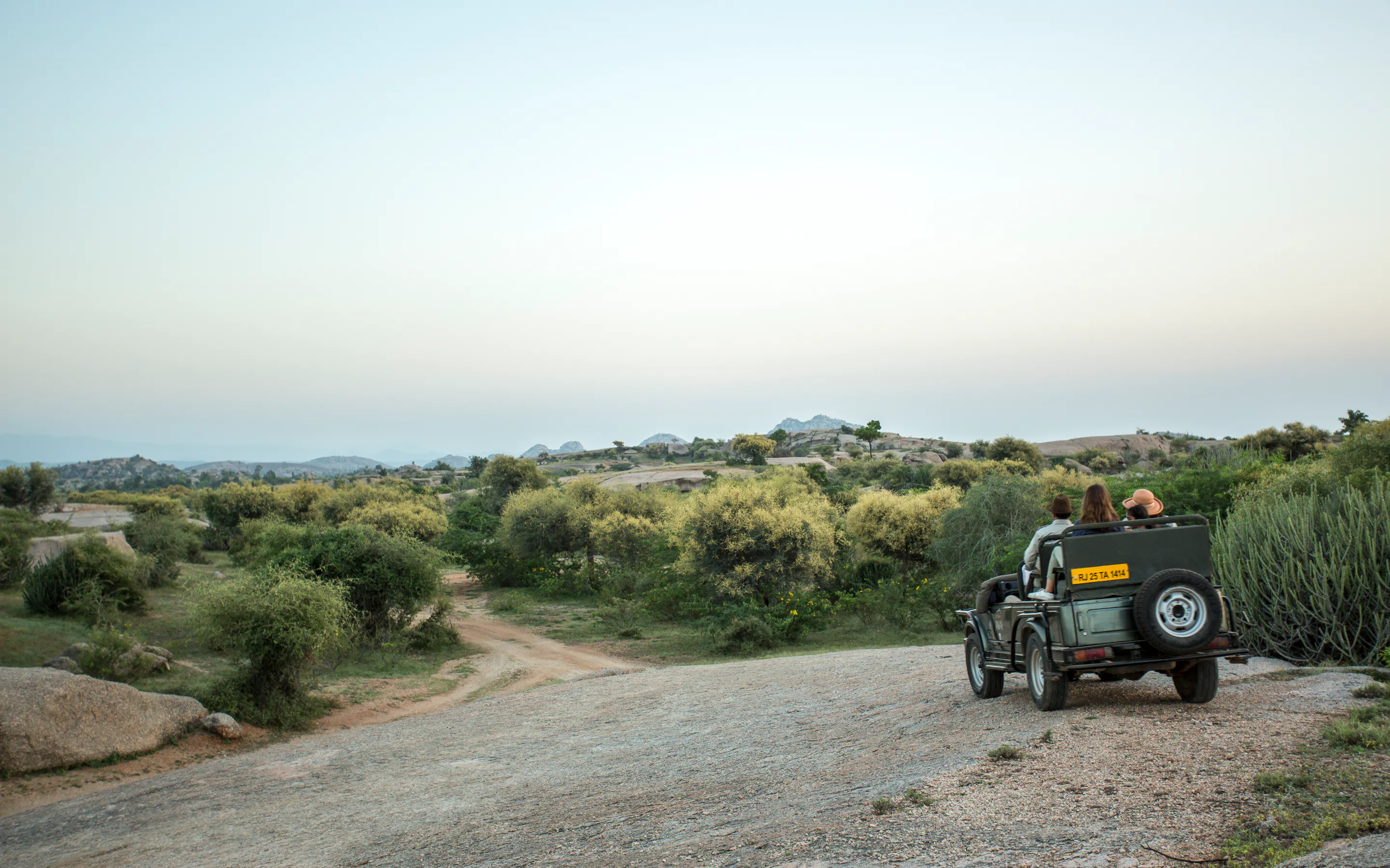 A safari jeep pauses on a gravel track above low scrub, with two guests seated inside and rounded hills beyond.