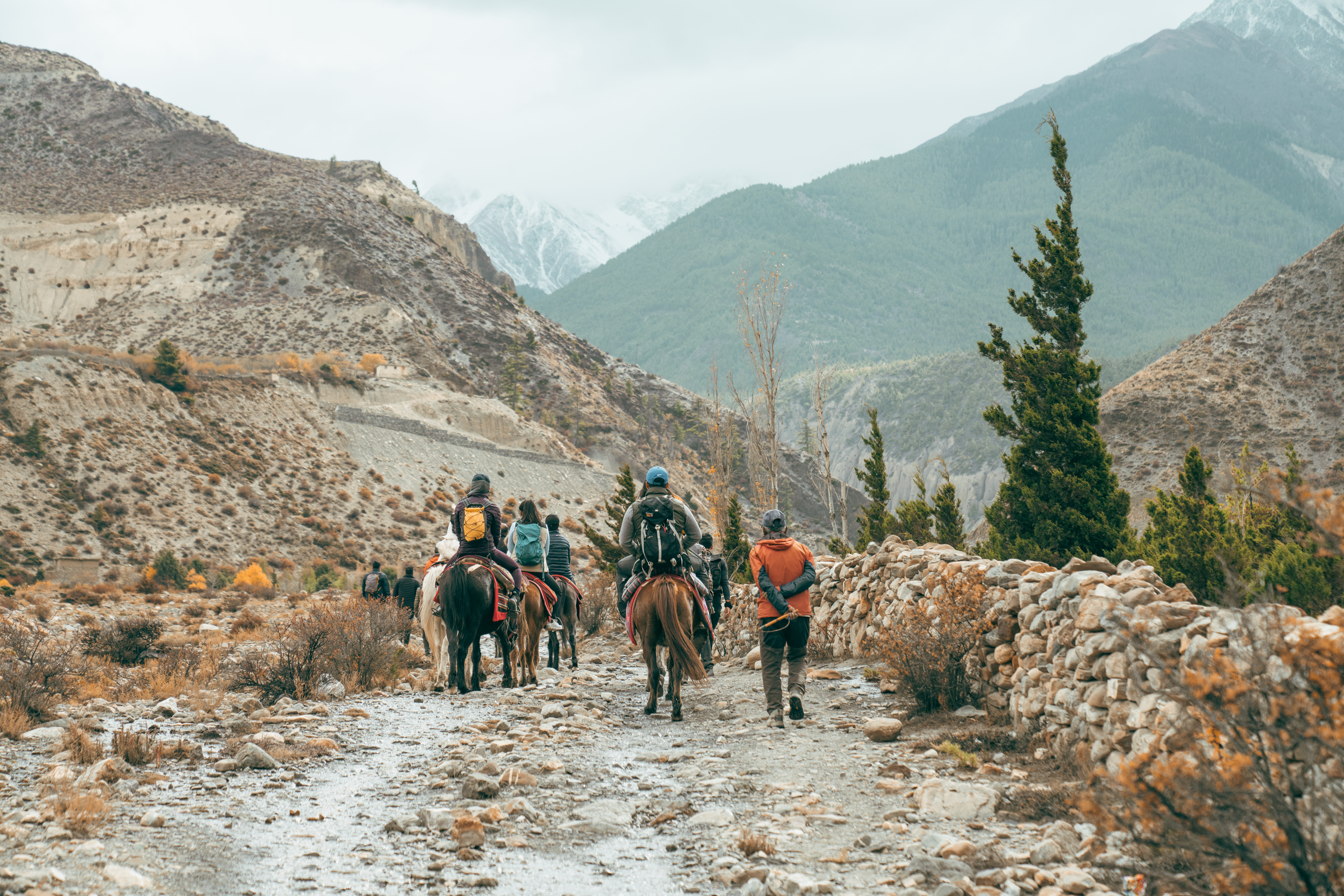 Riders and guides follow a rocky trail through Mustang, with stone walls, sparse trees, and mountain slopes ahead.