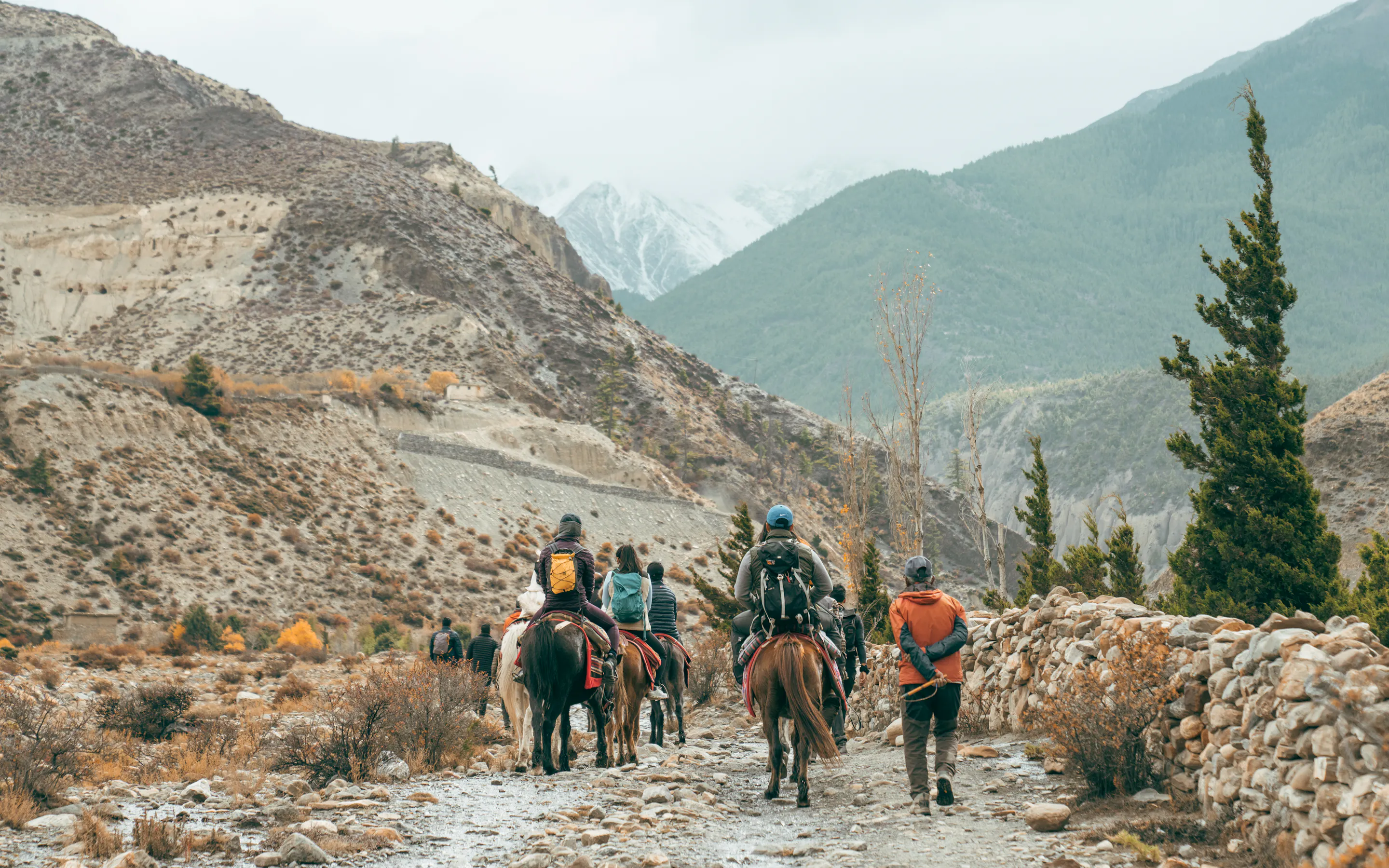 Riders and guides follow a rocky trail through Mustang, with stone walls, sparse trees, and mountain slopes ahead.